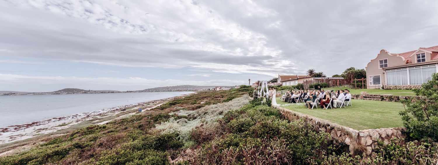 Outdoor coastal wedding ceremony with guests seated on green lawn overlooking a scenic beach and ocean under cloudy skies.