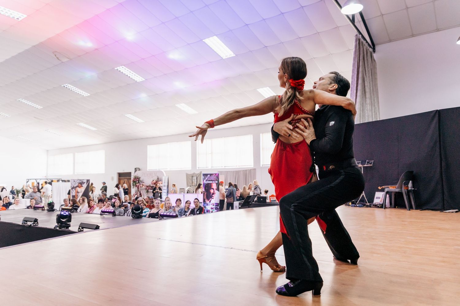 A couple performs a dramatic Latin dance routine in a bright studio, the woman in a red dress dipping gracefully.