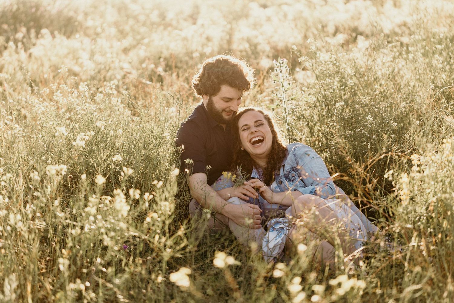 Couple sits together in golden wildflower field during sunset photoshoot.