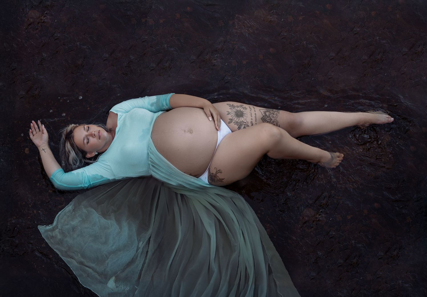 A pregnant figure in a flowing turquoise dress lies gracefully on a dark background during a maternity photoshoot.