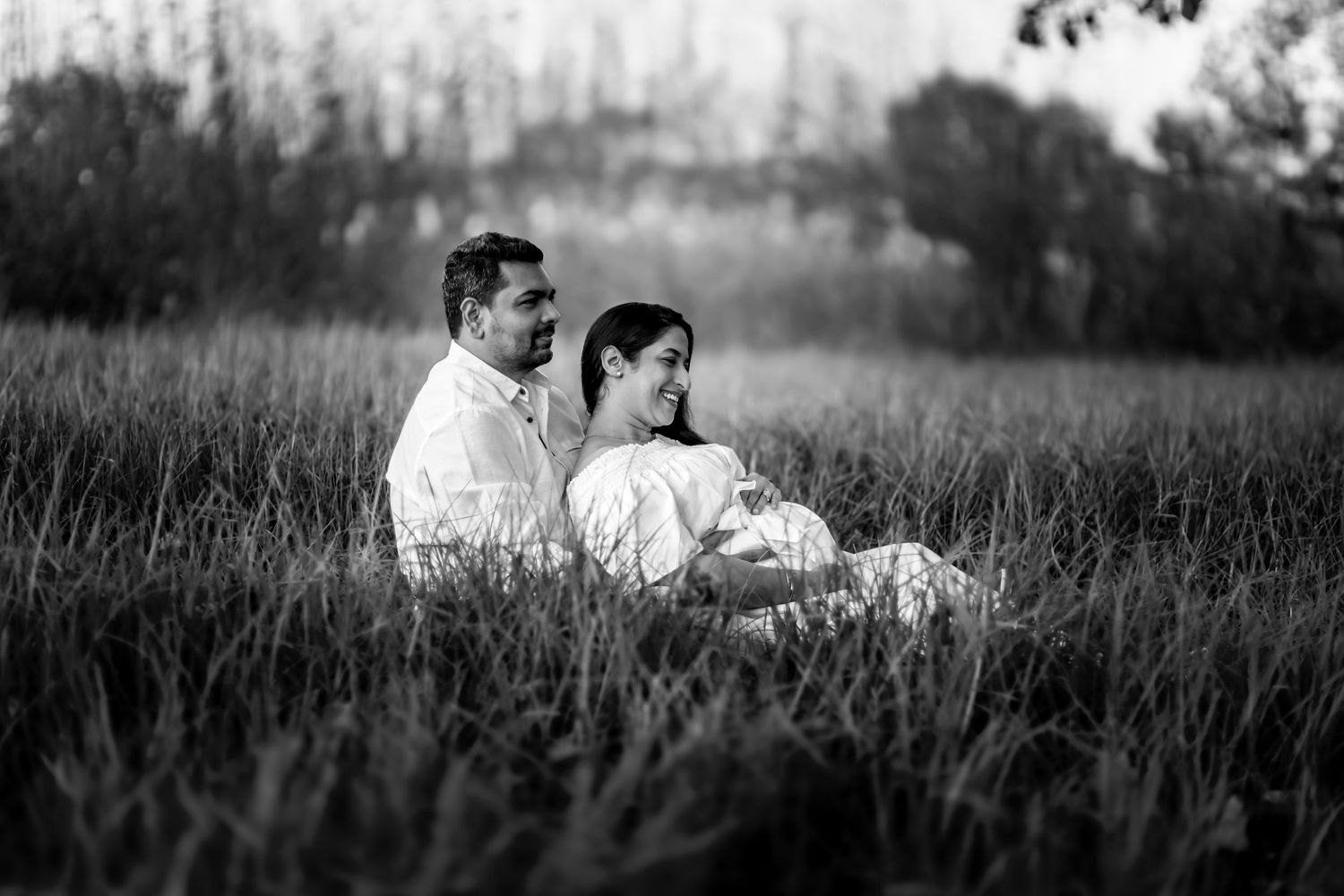 A romantic couple sits together in a grassy meadow during sunset creating a silhouetted scene.