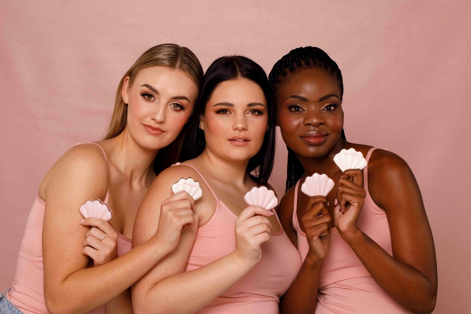 Three women in pink dresses pose together while holding pink cupcakes against a pink background.