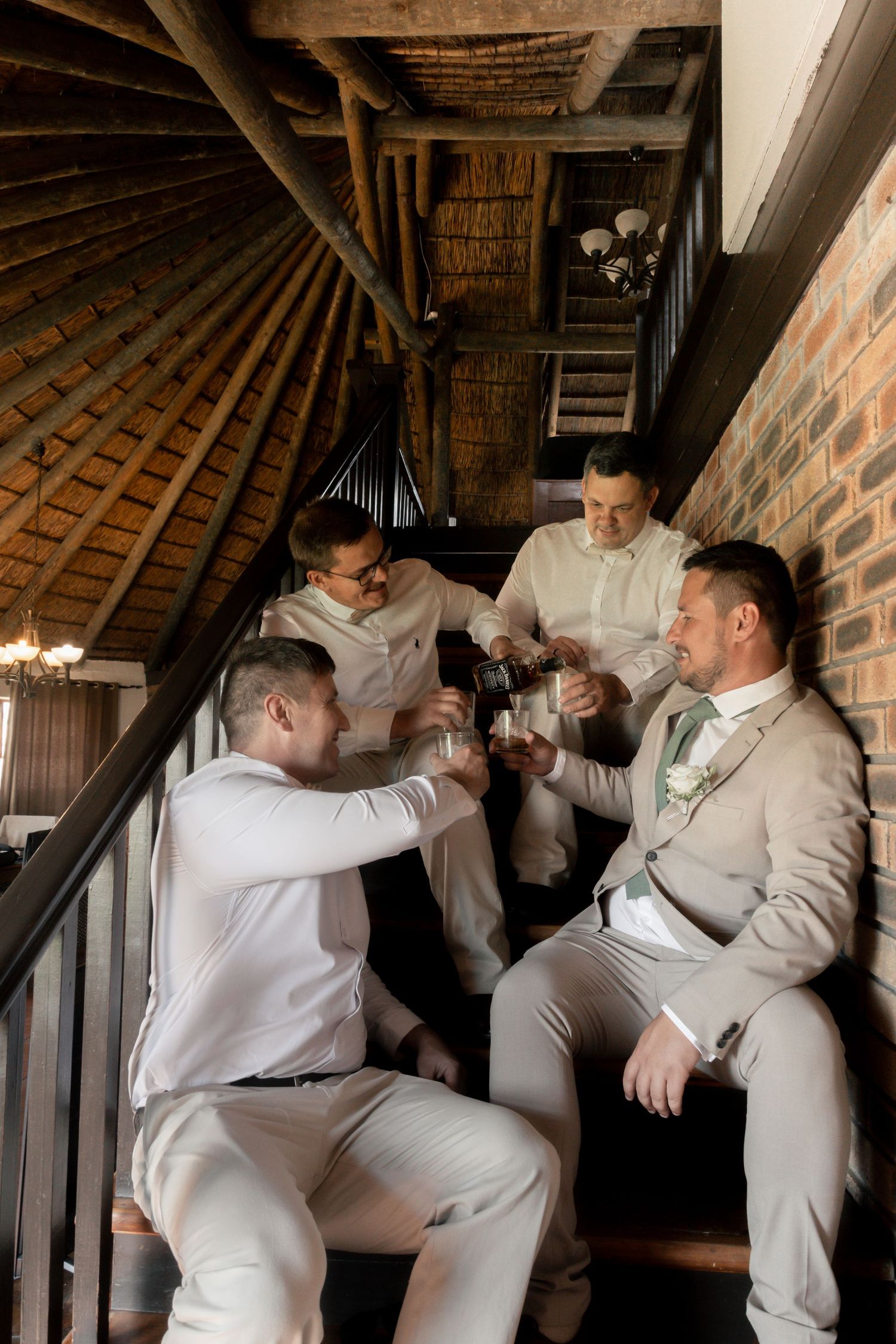 Four groomsmen in light suits toasting drinks on rustic wooden stairs in a thatched venue before a wedding ceremony.