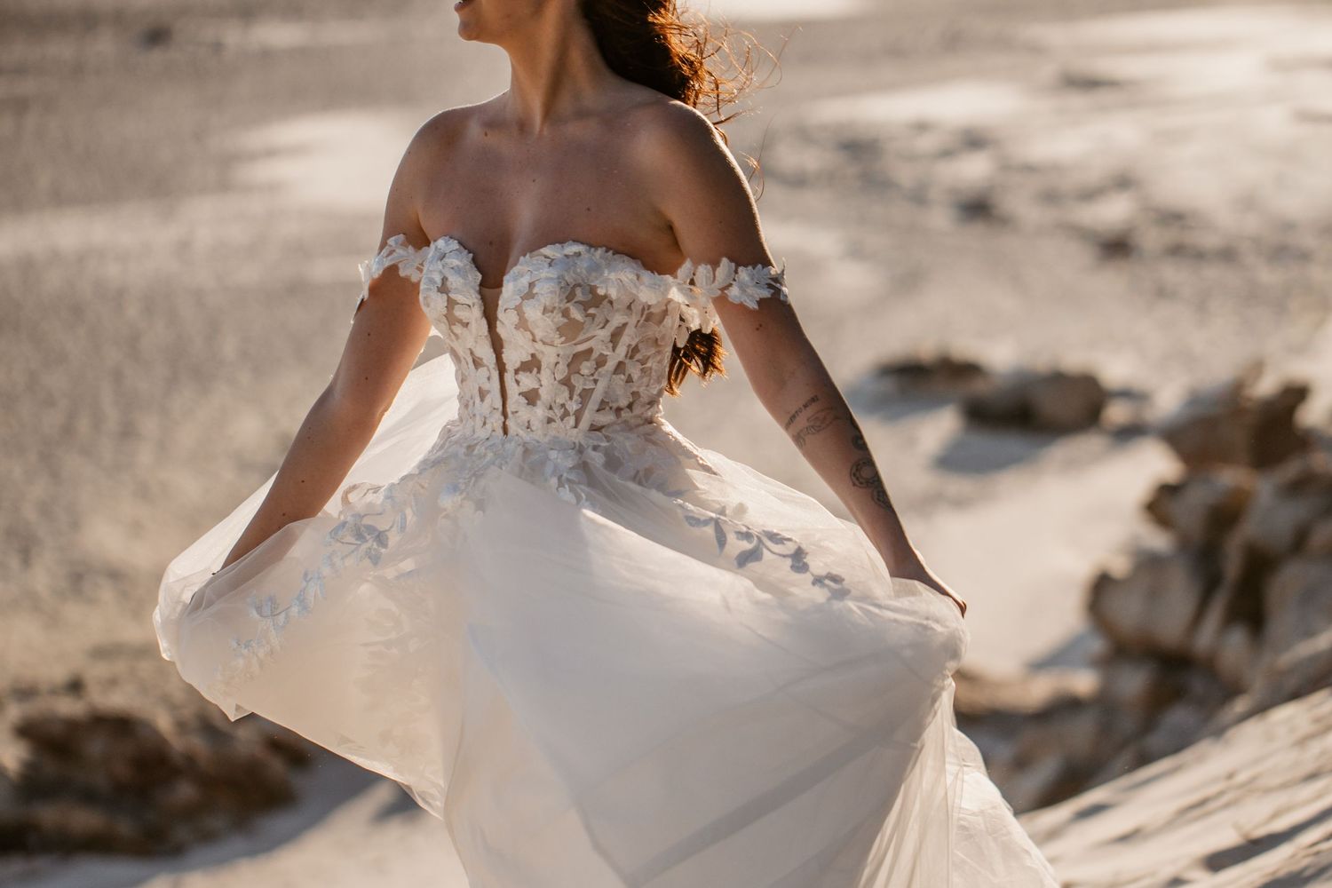 Bride in an off-shoulder lace wedding gown with floral details, flowing skirt, posing on a sandy beach.