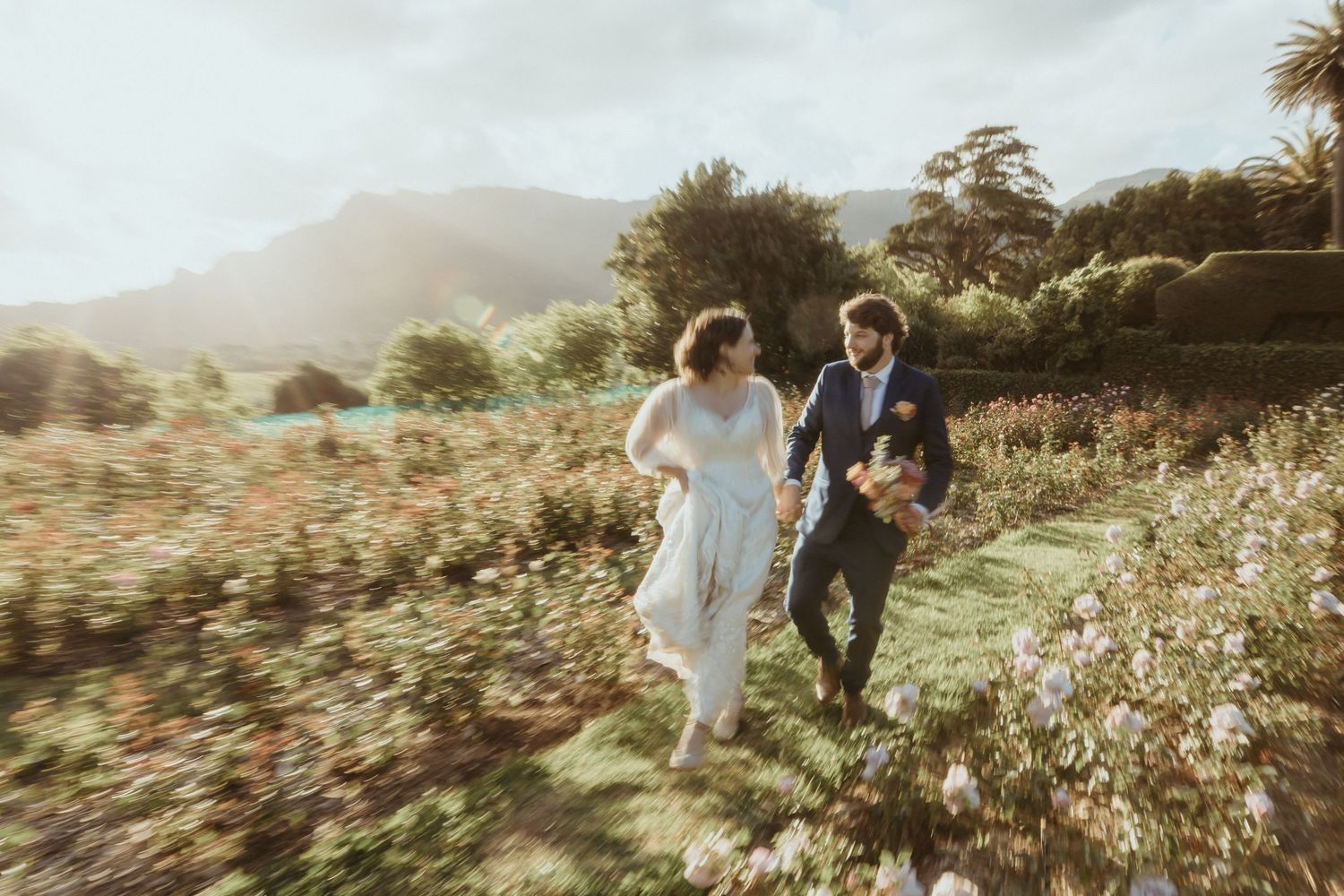 Bride and groom running through a lush garden with mountains in the background on their wedding day.