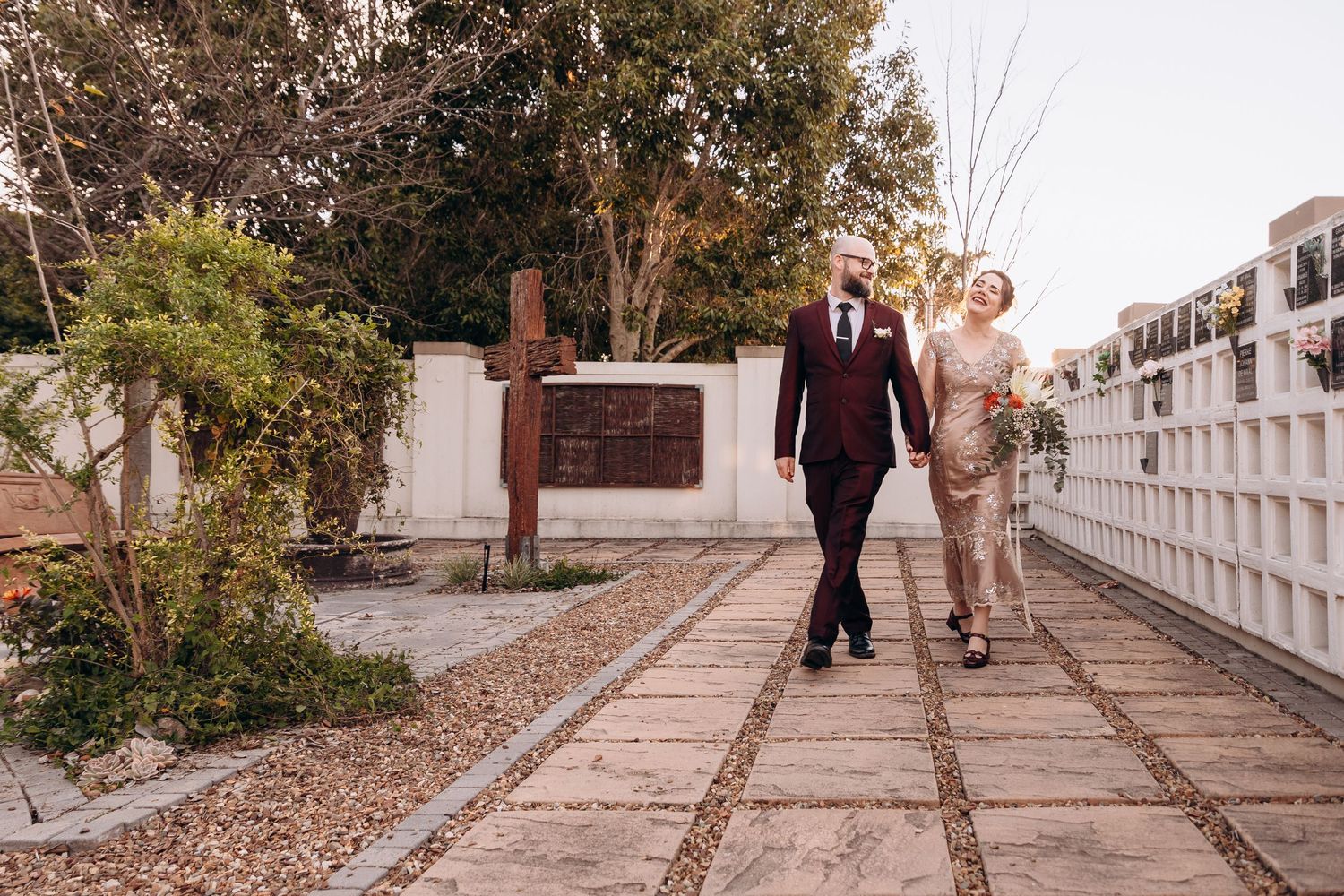 A couple in formal wedding attire walks hand-in-hand along a paved garden path surrounded by lush greenery.