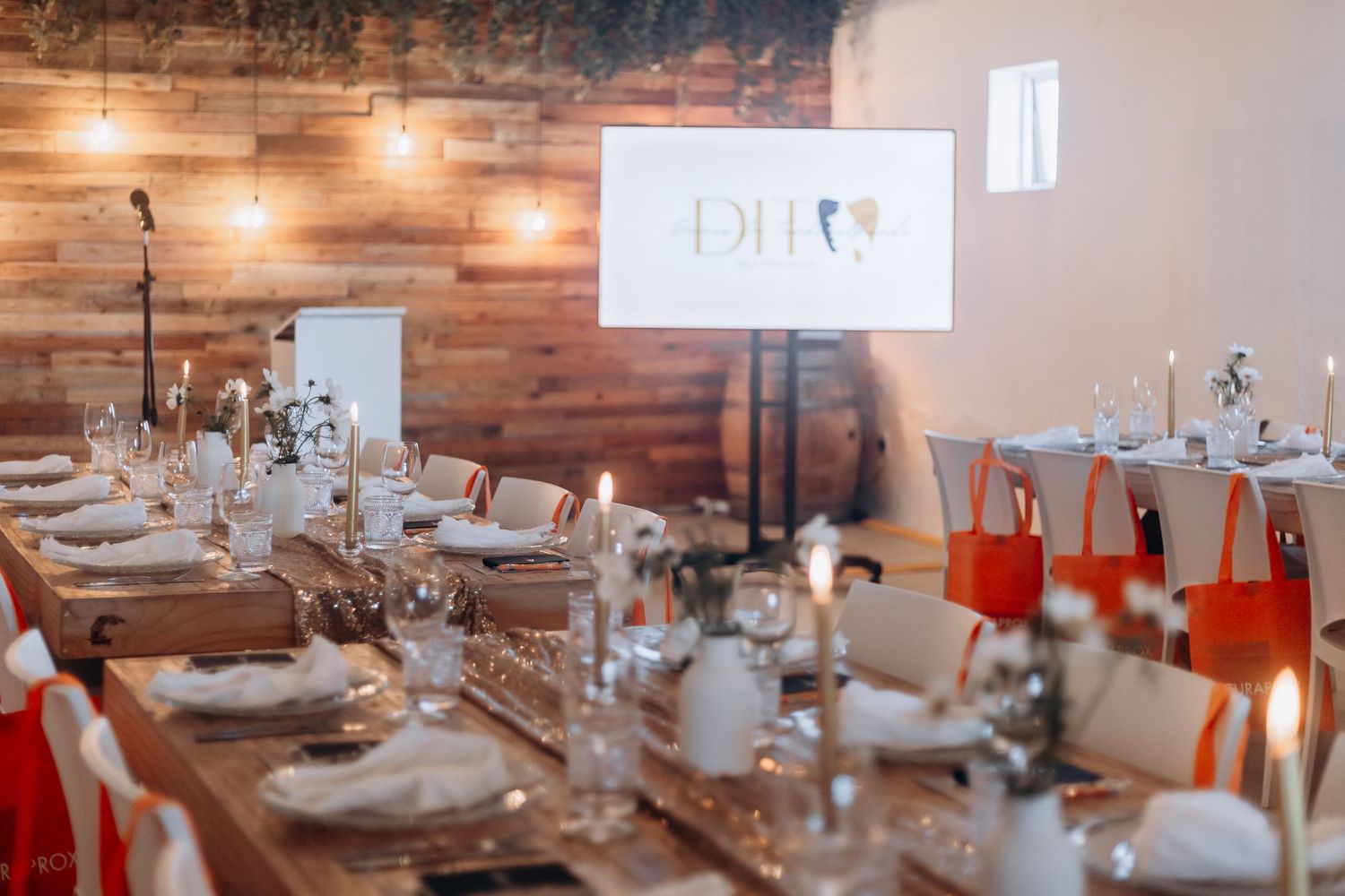 Elegant event dining room with long wooden tables set with candles, glassware, and red gift bags under warm string lights.