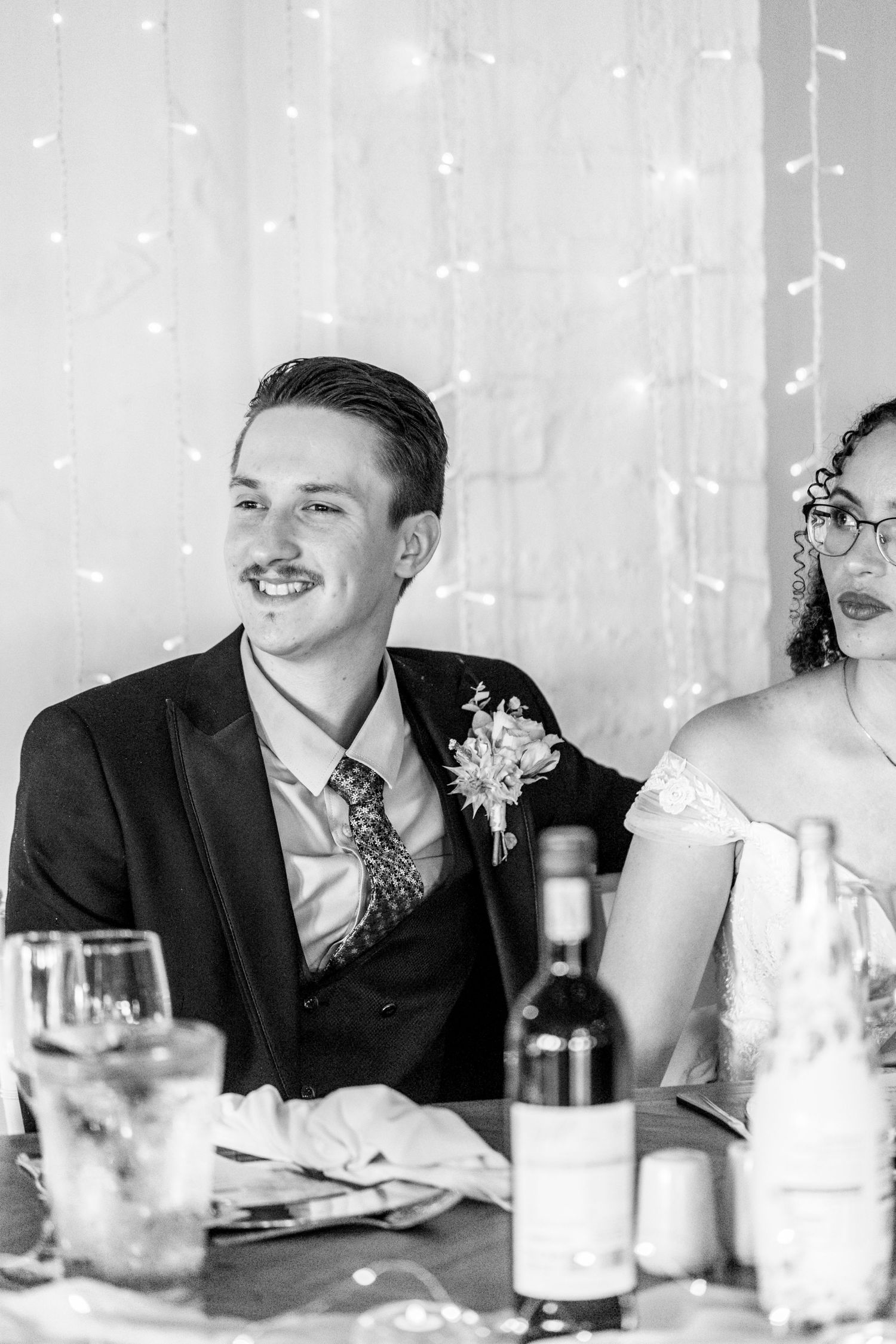 Black and white photo of wedding guests laughing and enjoying themselves at a reception table with drinks.