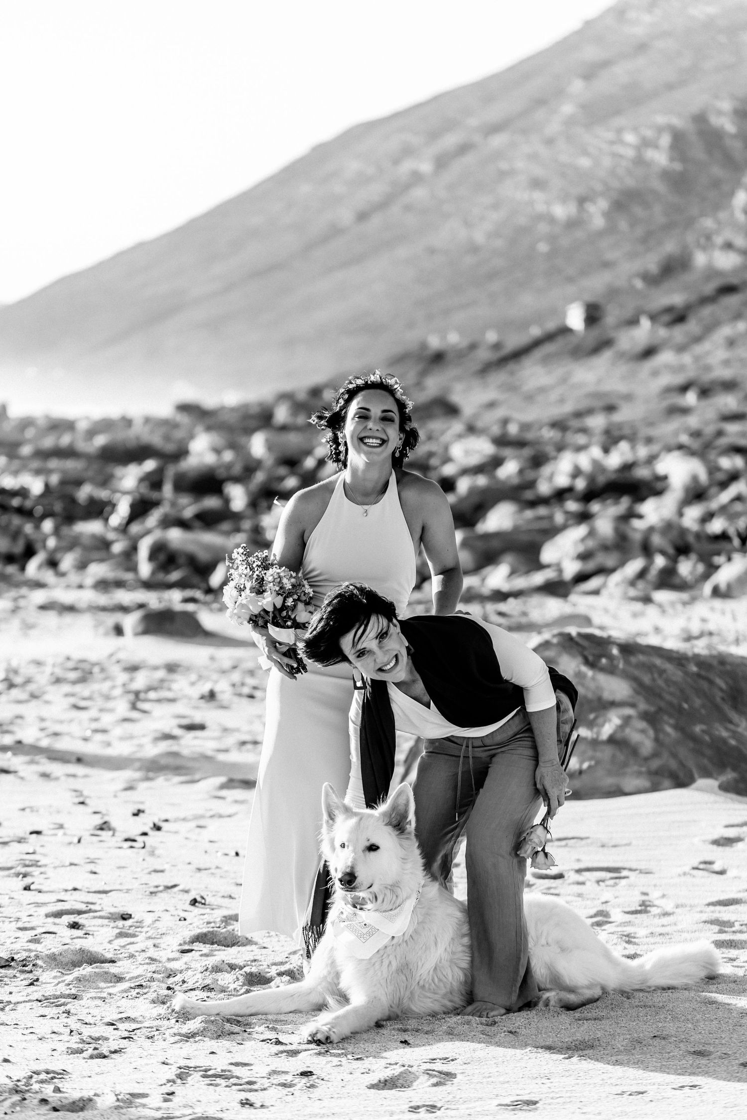 A joyful moment captured in black and white on a beach with two dogs and a bridal bouquet against mountainous backdrop.