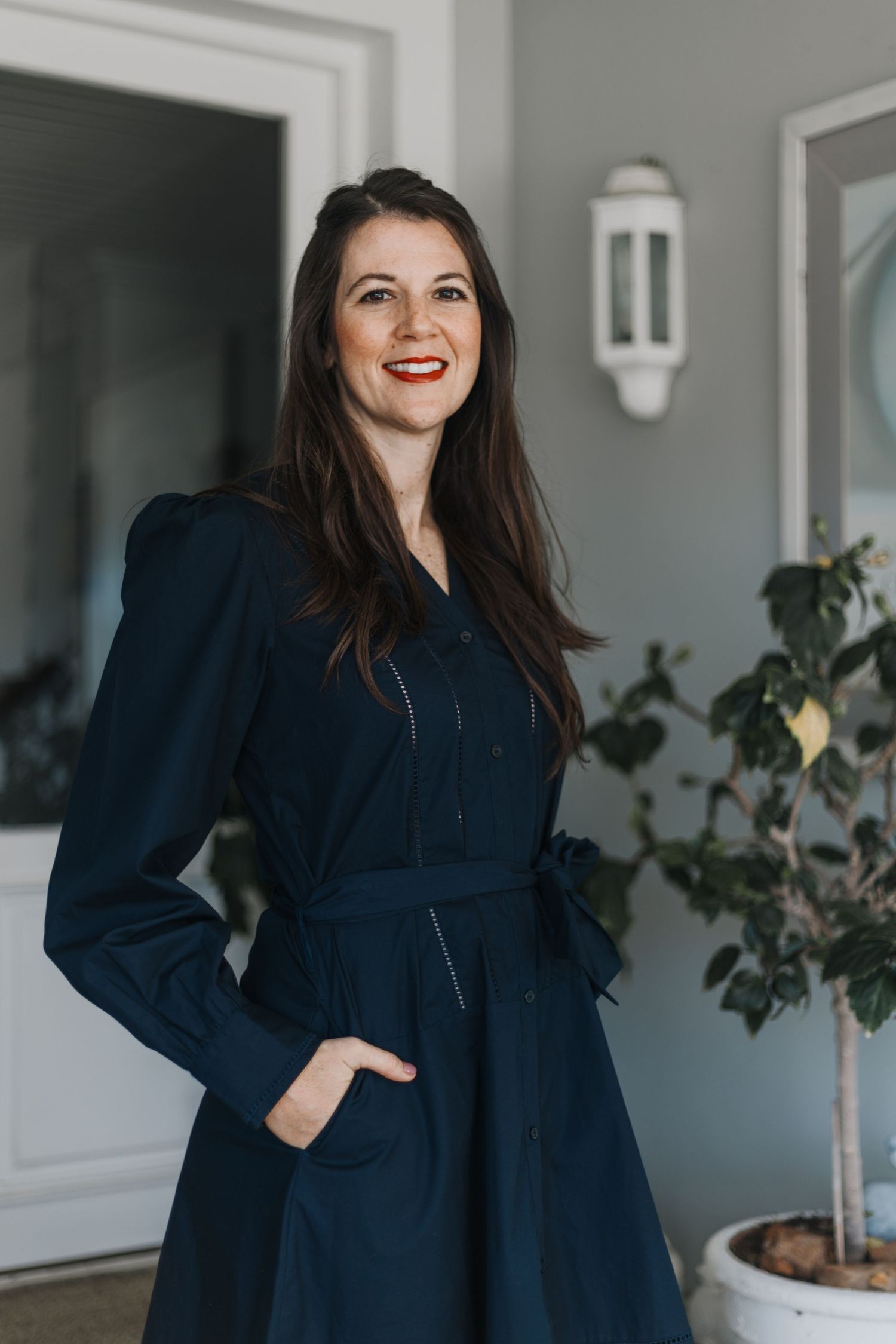 Stylish navy blue button-down dress with long sleeves and belt worn in front of white wall with decorative lantern.