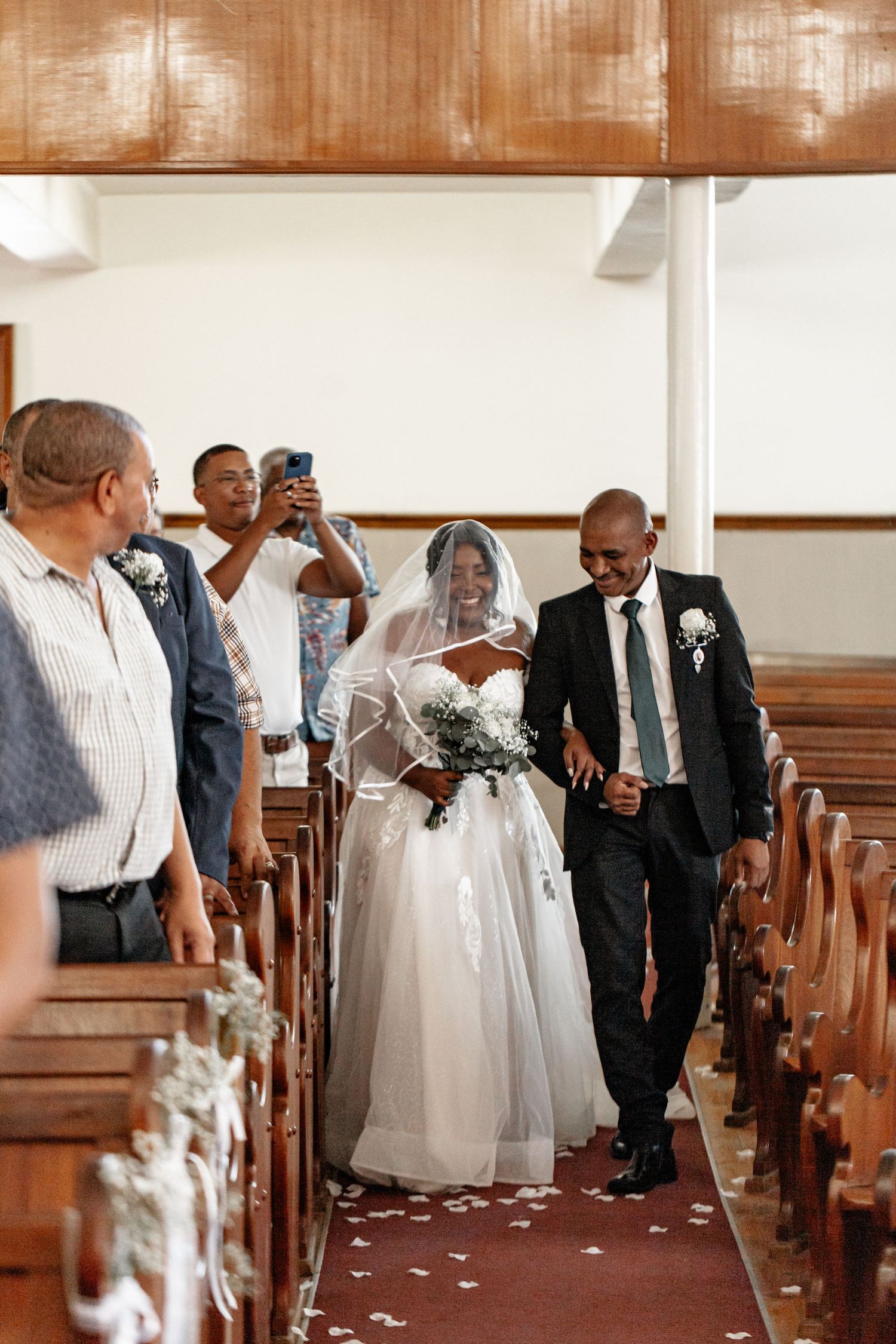 Guests walk down a church aisle decorated with white petals while taking photos during a wedding ceremony.