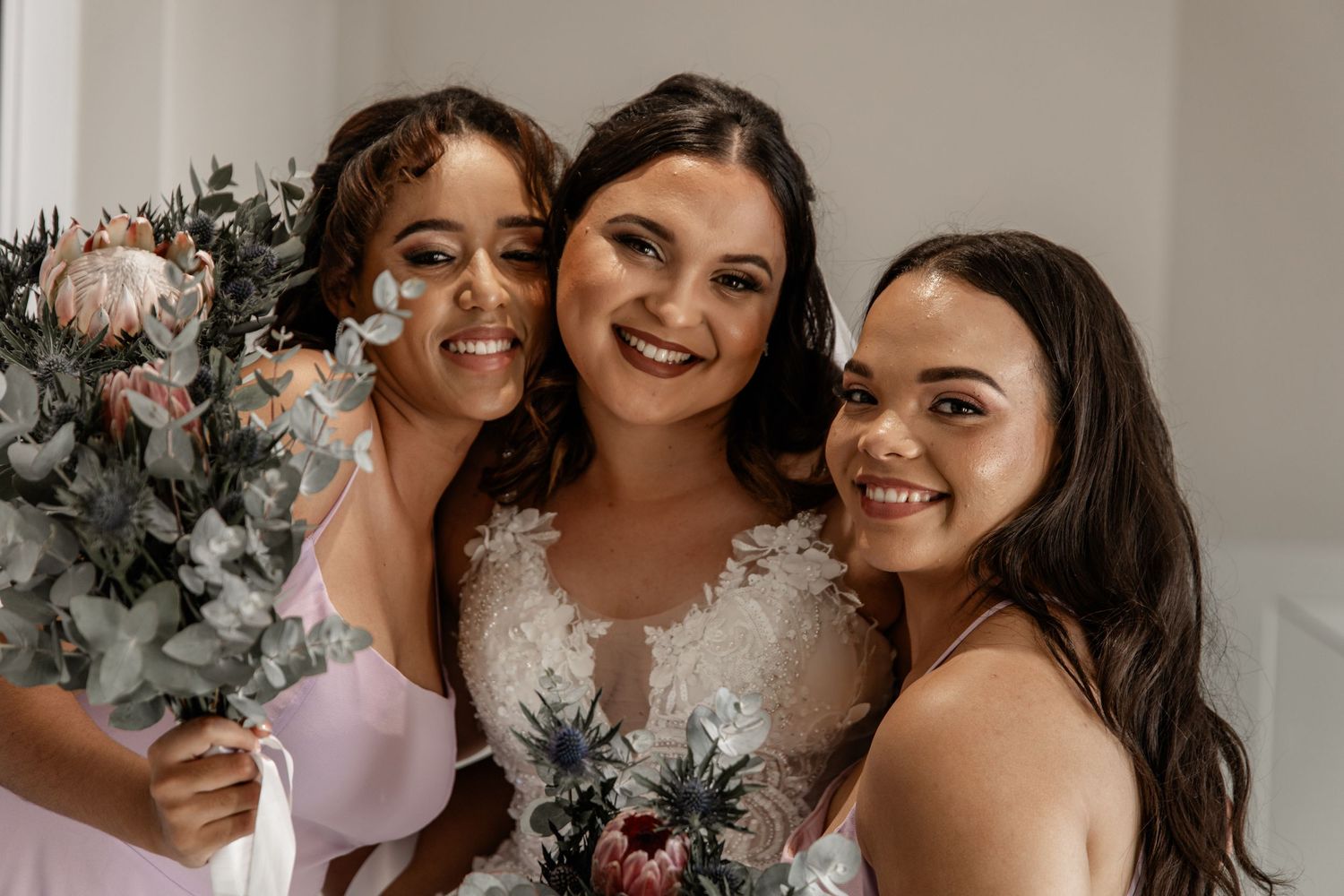Three people in wedding attire smile together with floral decorations in the background.
