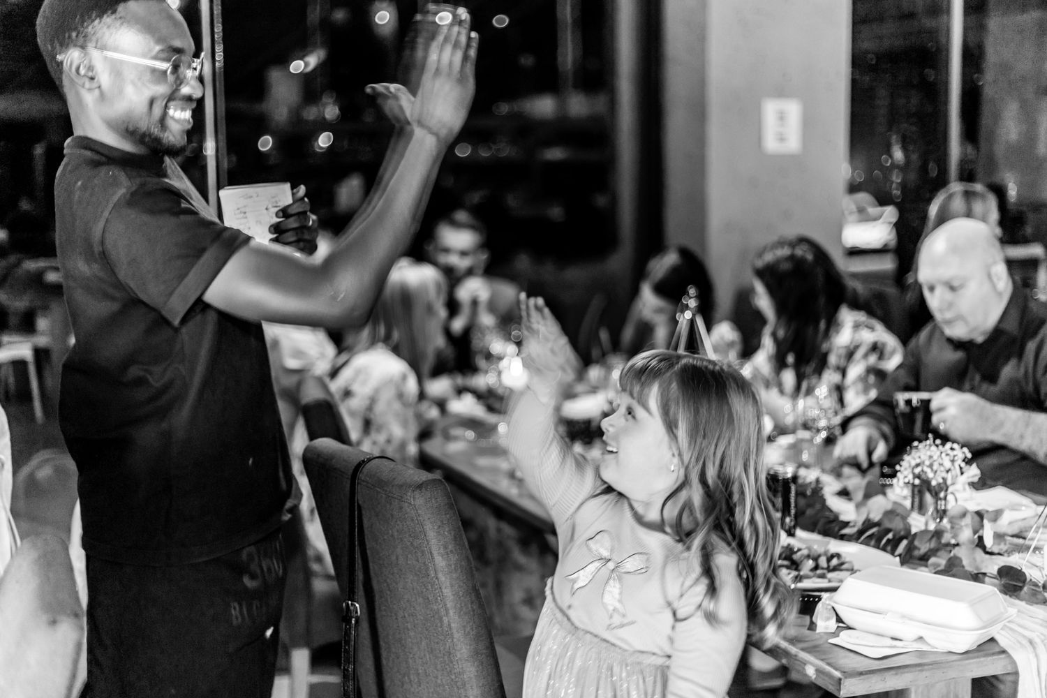 A black and white photo shows a young child standing and singing with enthusiasm during what appears to be a restaurant gathering.