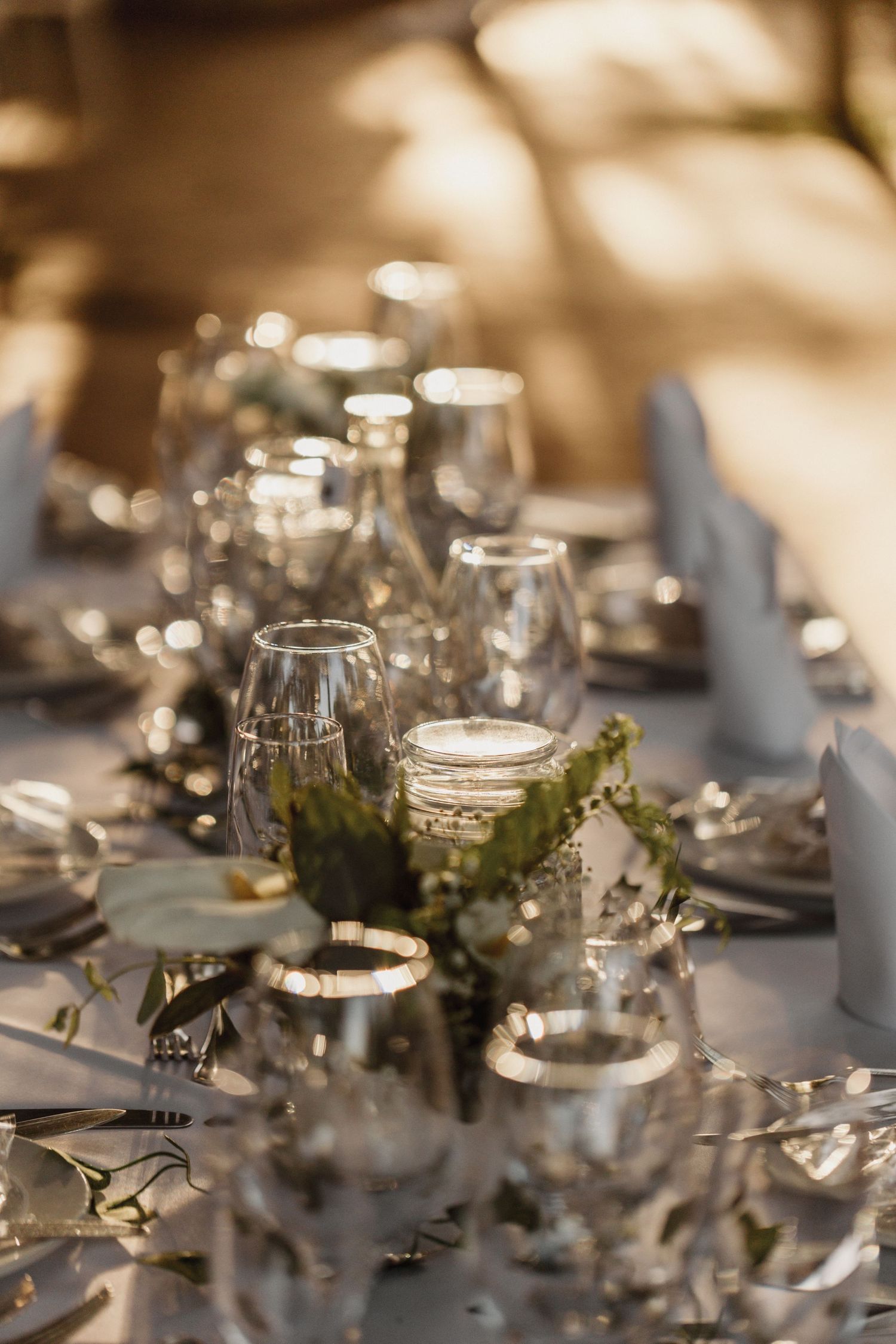 Elegant table setting with wine glasses and mason jars decorated with greenery in soft, warm lighting.