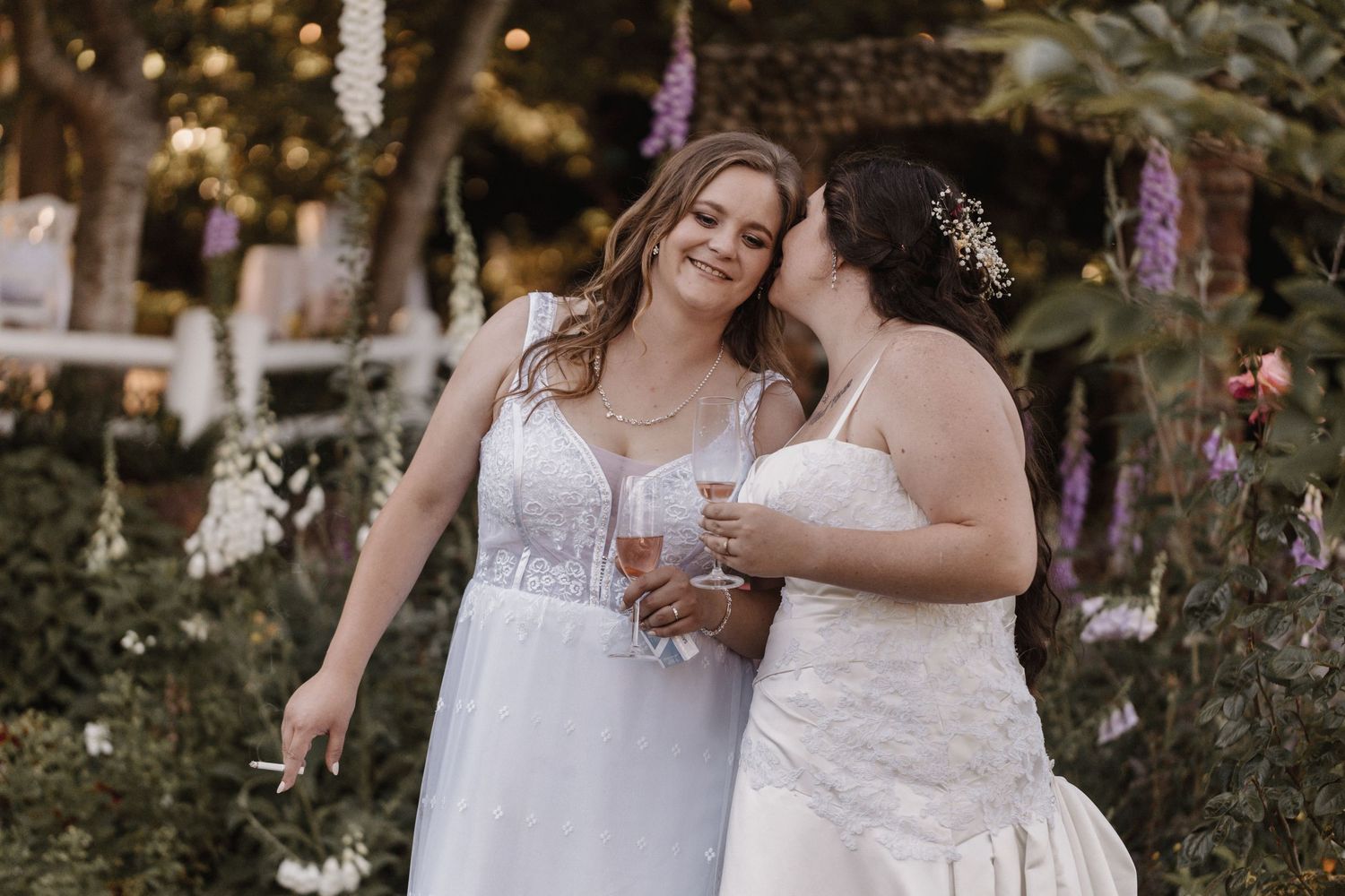 Two people in white wedding dresses share a laugh while holding glasses during an outdoor garden celebration.