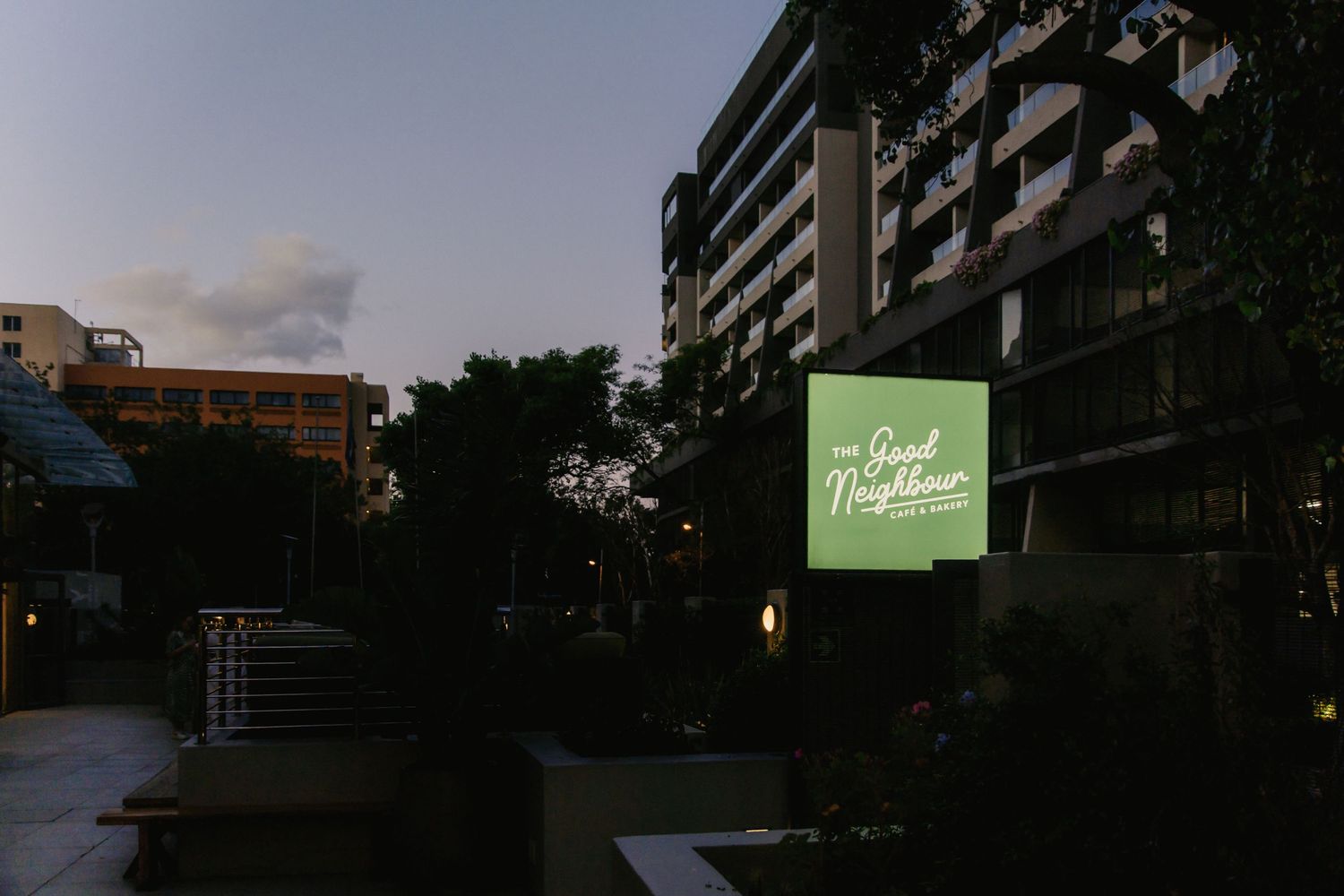Glowing green neon sign illuminates a dark urban street corner at dusk.