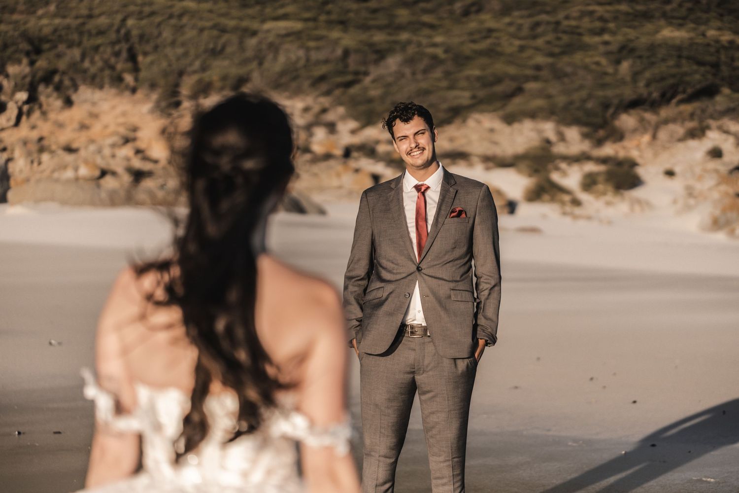 Groom in gray suit smiling at bride on sandy beach during outdoor wedding ceremony.