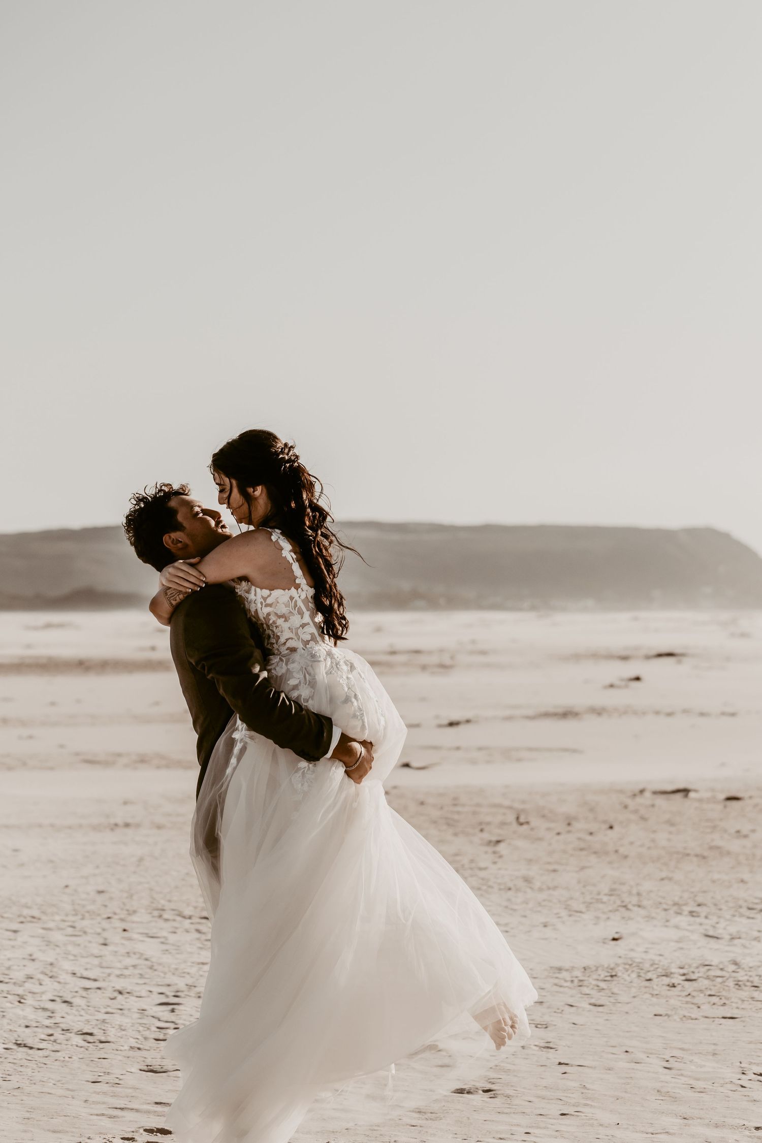 A couple embraces on a sandy beach, the bride in a flowing white lace gown, her veil blowing in the wind.