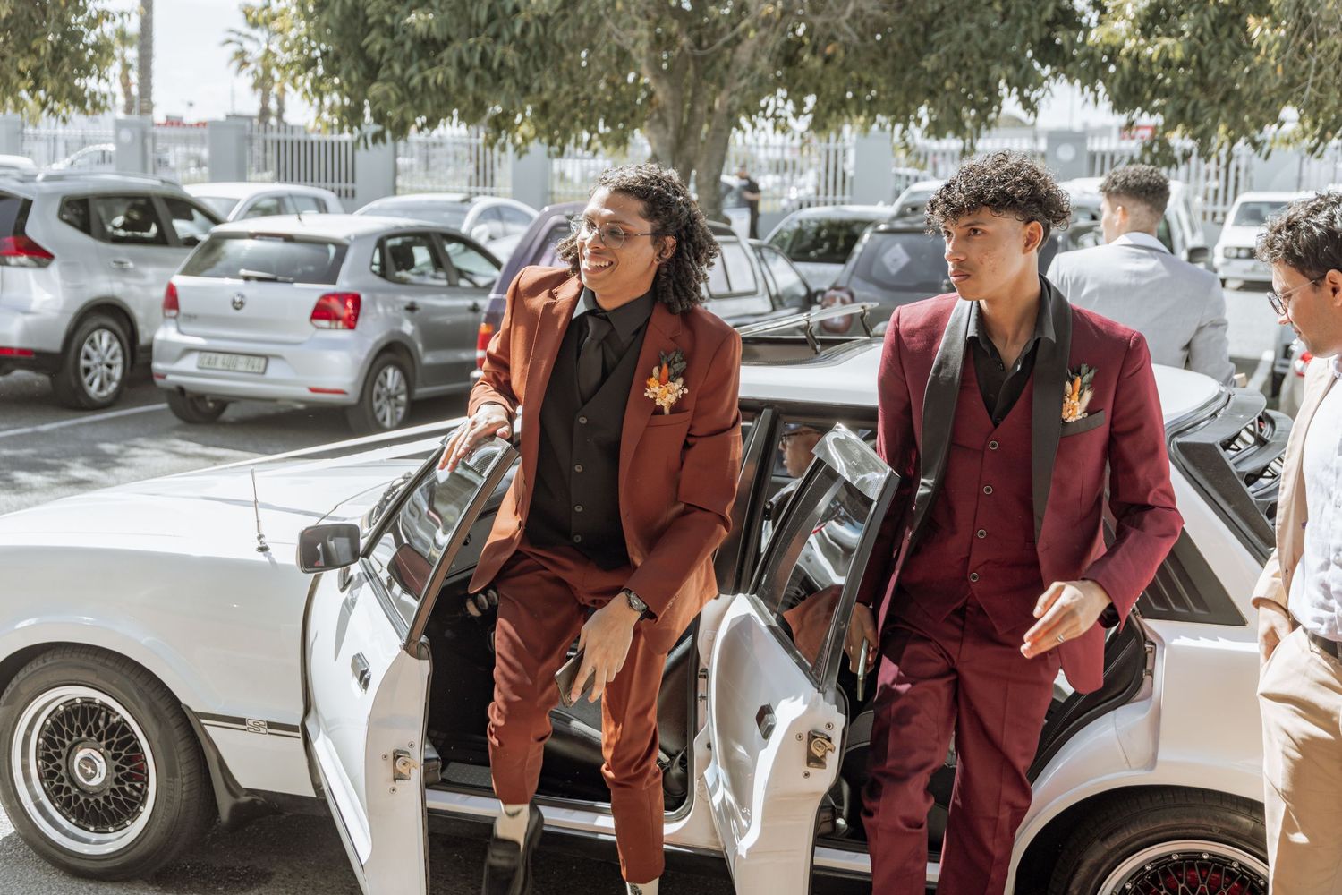 Two people wearing matching burgundy suits exit a white classic car in a parking lot.