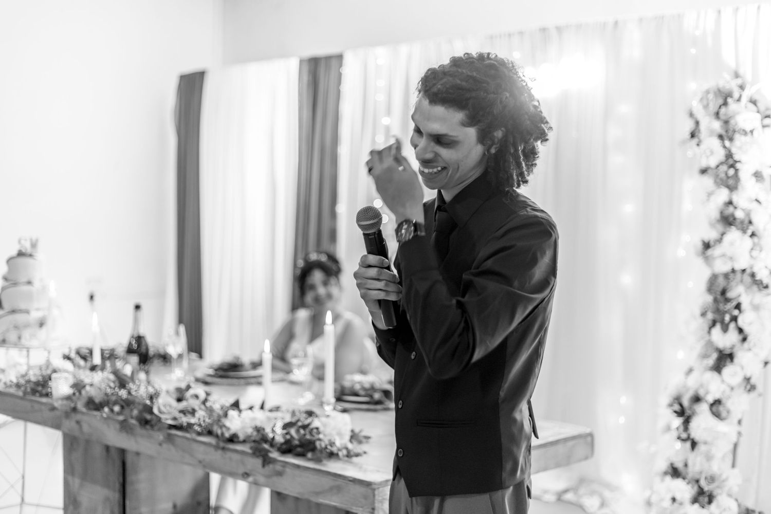 Someone delivers an emotional speech at a wedding reception with floral decorations and white curtains in the background.