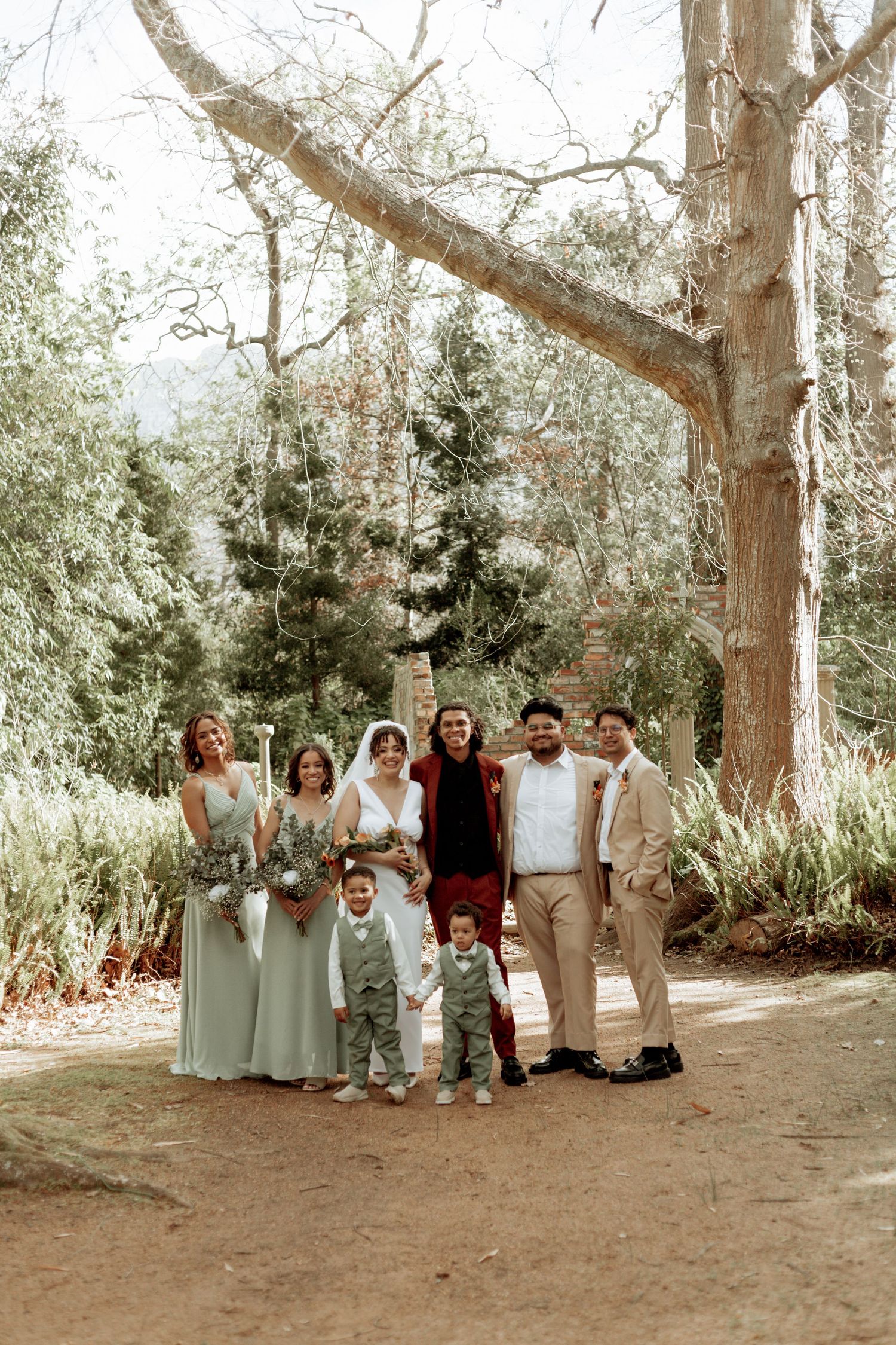 Wedding party in sage green and beige attire poses under tall trees on a forest path.