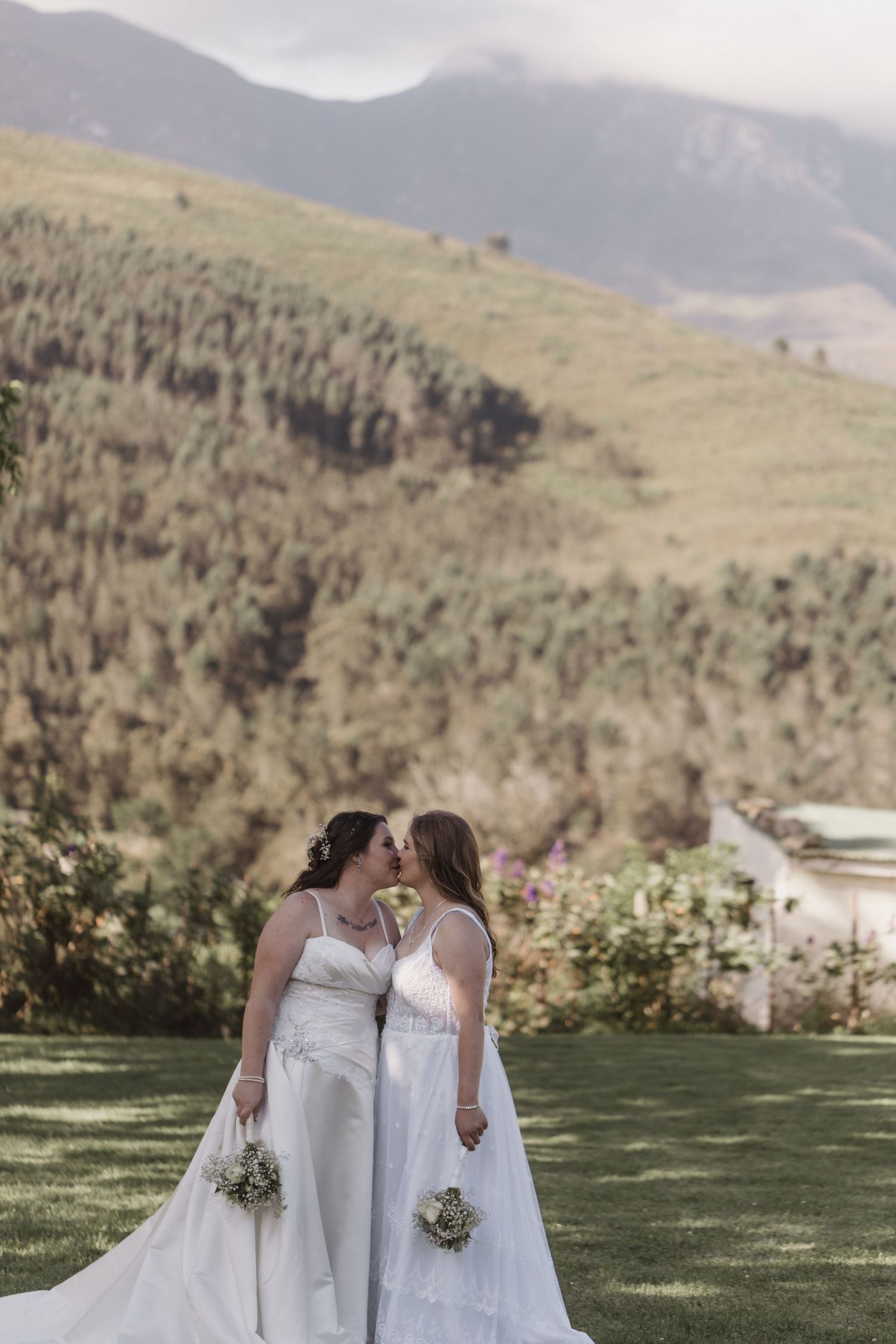 Two brides share a tender moment on a grassy hilltop with mountains in the background on their wedding day.