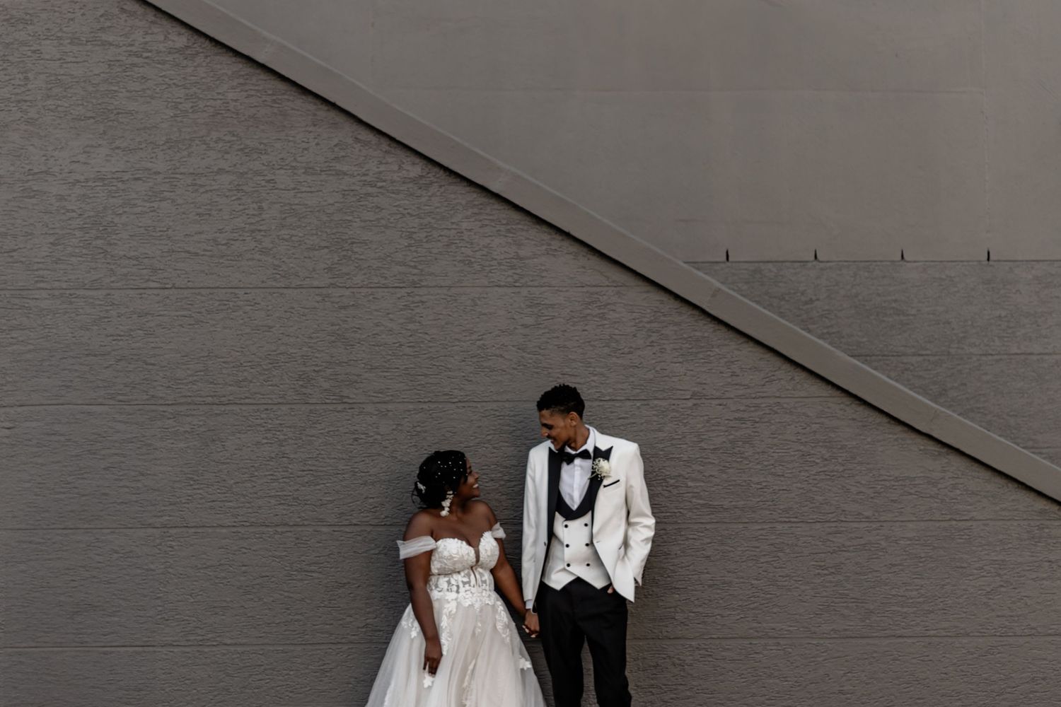 A couple in wedding attire walk together on gray textured concrete during an outdoor photo session.