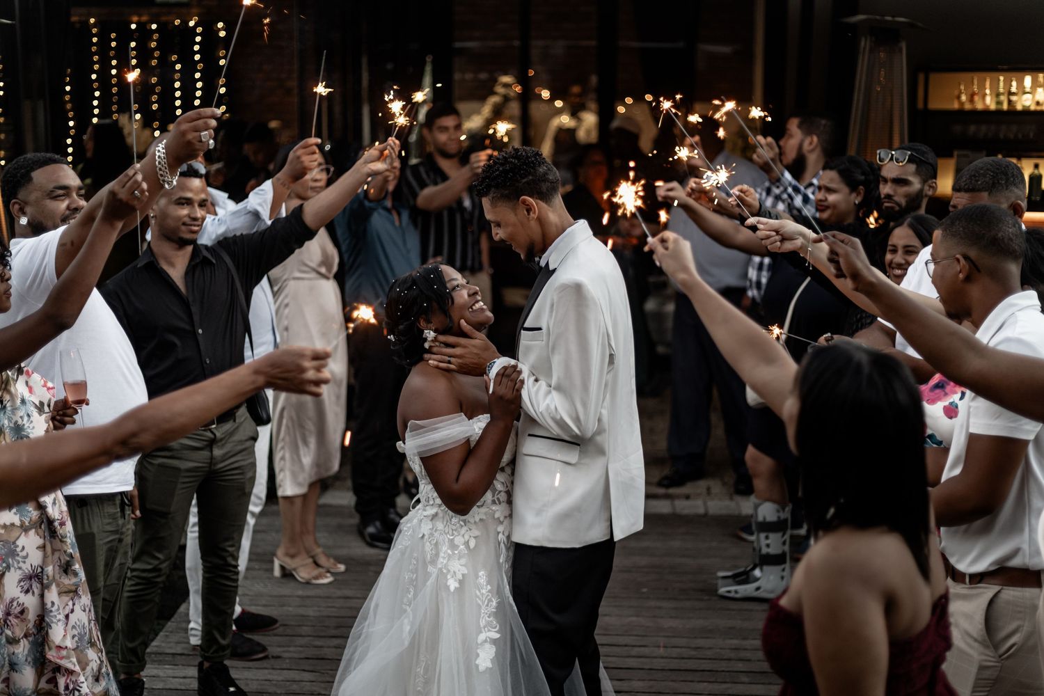 Wedding guests dance and celebrate at an indoor reception under ambient lighting with hands raised in joy.