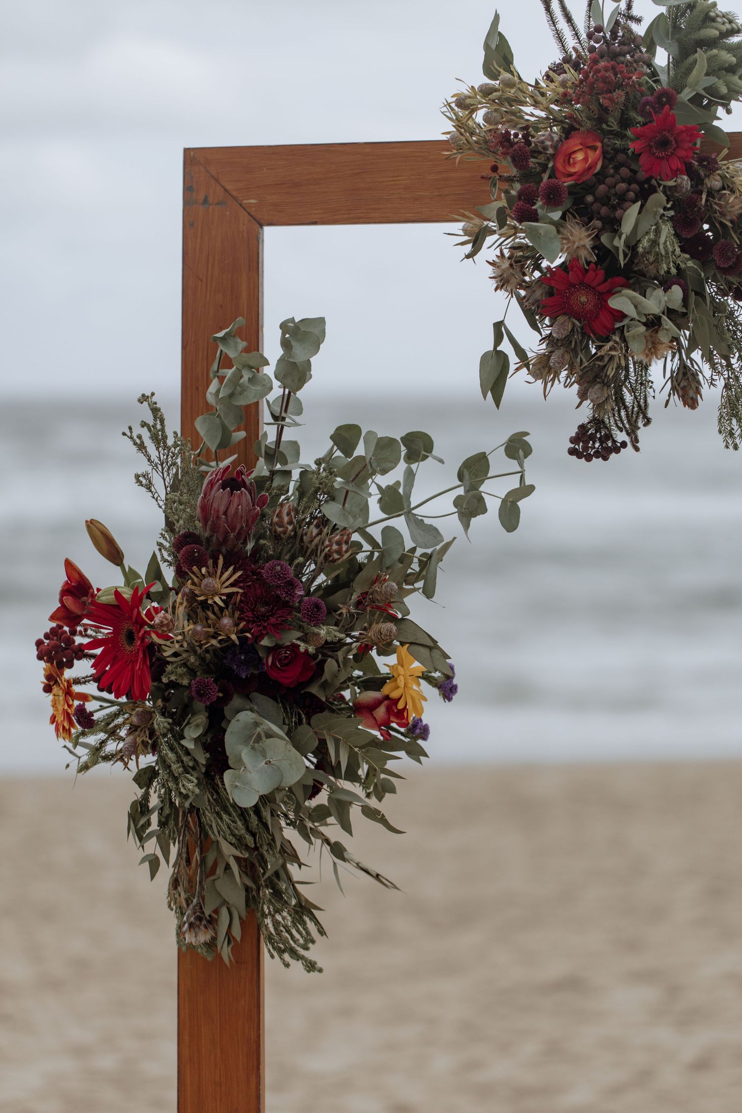 Wooden wedding arch decorated with deep red and burgundy floral arrangements on a misty beach backdrop.