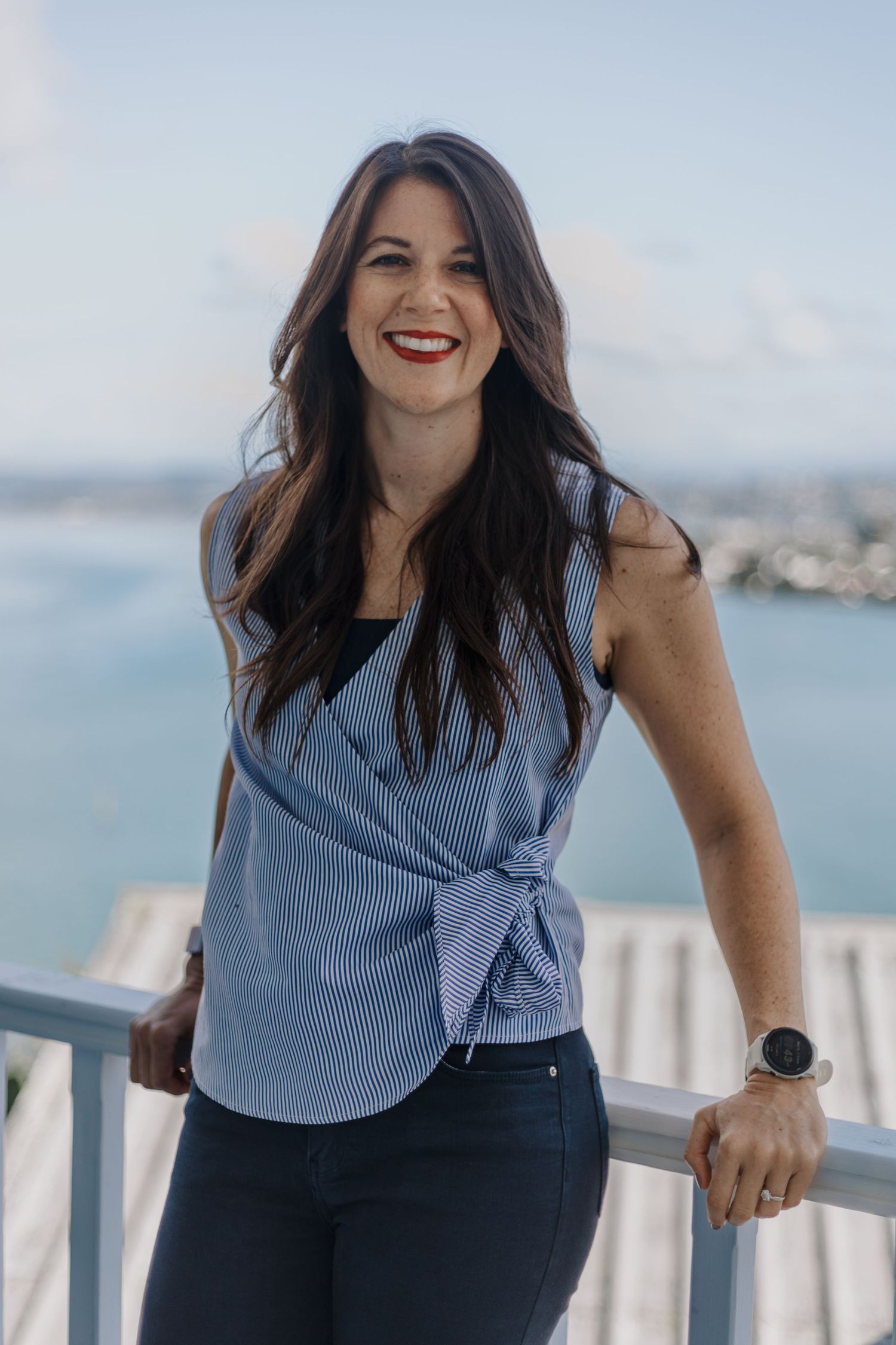Woman in striped blue top and dark pants poses outdoors on balcony with ocean view in background.