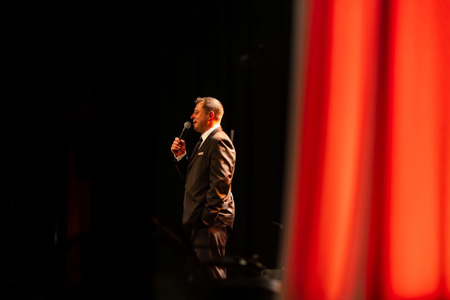 Man in suit speaking into microphone on stage with red curtain backdrop at a formal event.