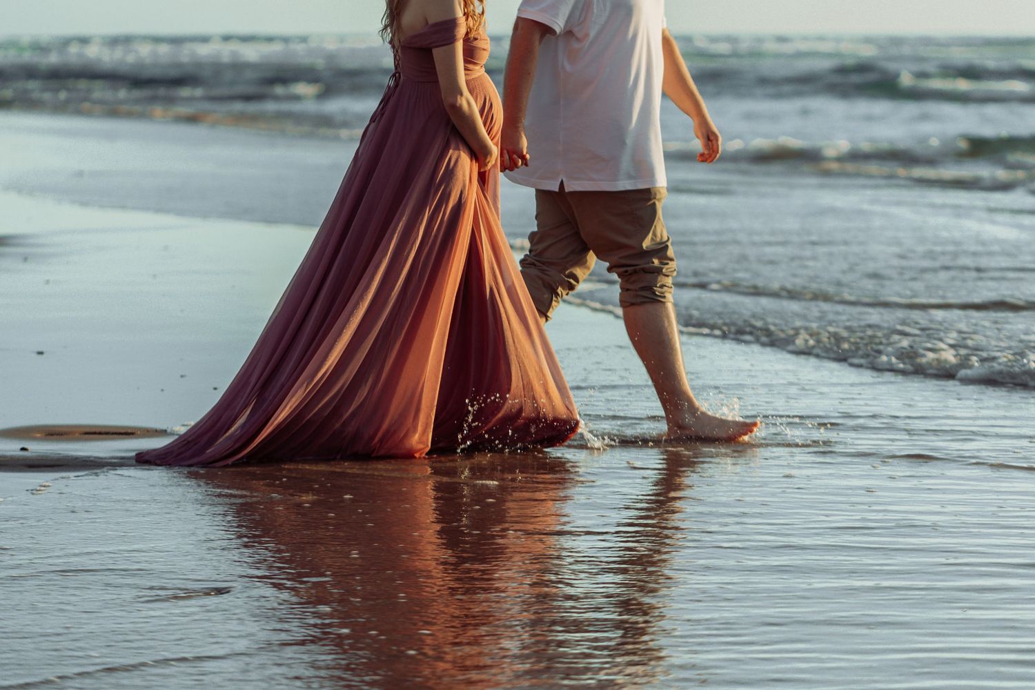 Bare feet walking along wet beach sand at sunset with long flowing dress skimming the water's surface.