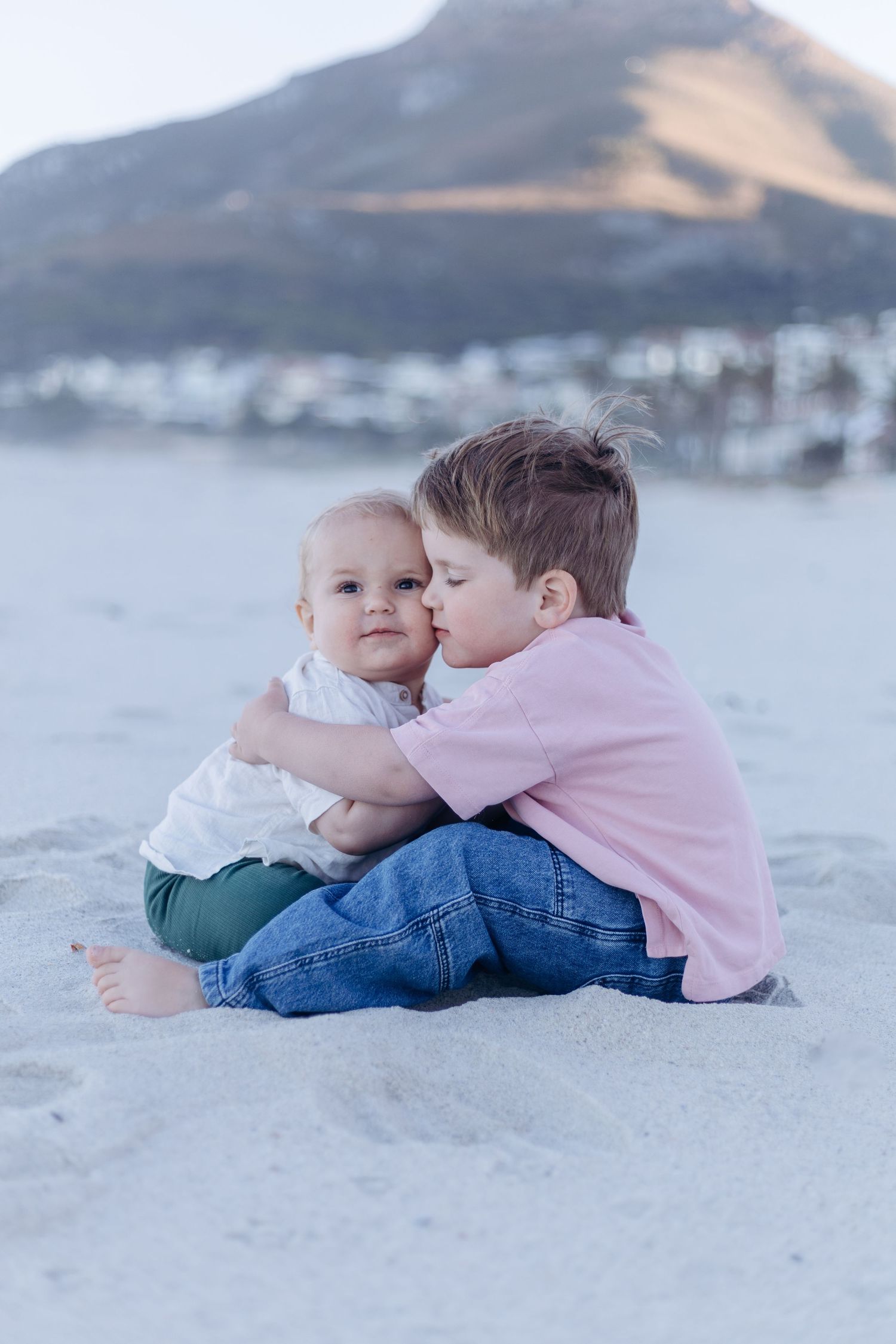 Two children lying on sandy beach in soft lighting with mountains in background.