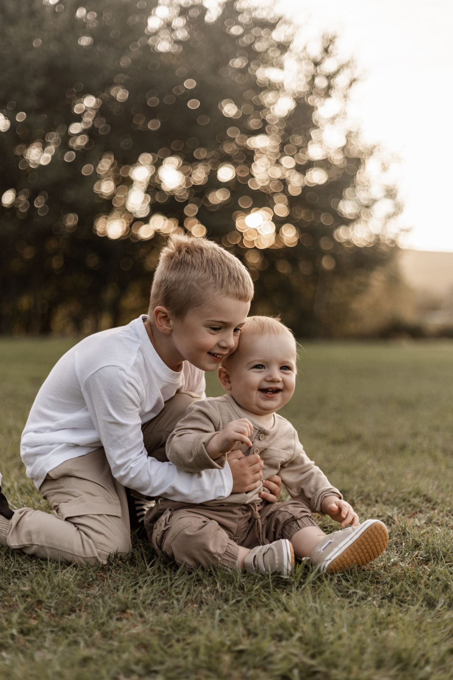 Two children in white and beige outfits share a sweet moment while sitting together on grass at sunset.