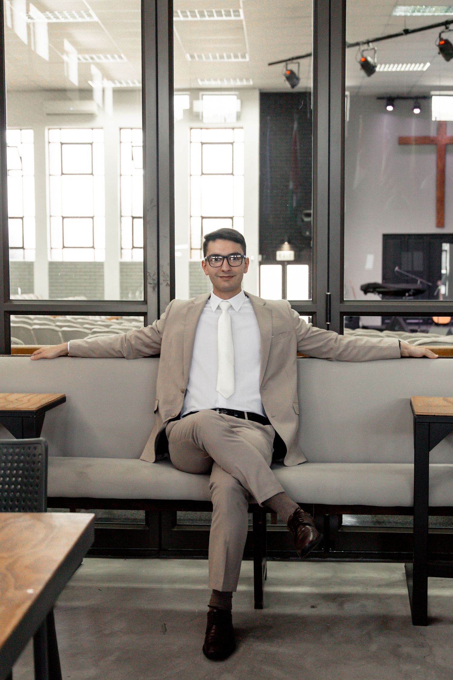 A person in a beige suit and glasses sits on a modern bench in an office space with glass partitions.
