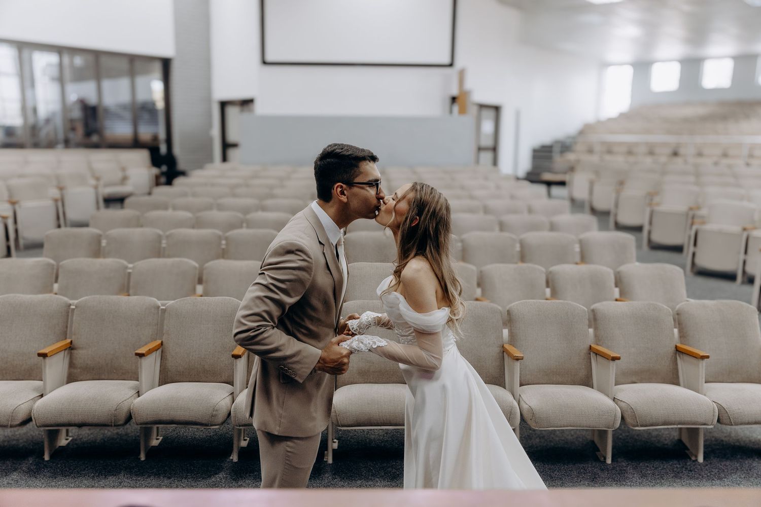 A couple shares a romantic moment in an empty church auditorium with white chairs.