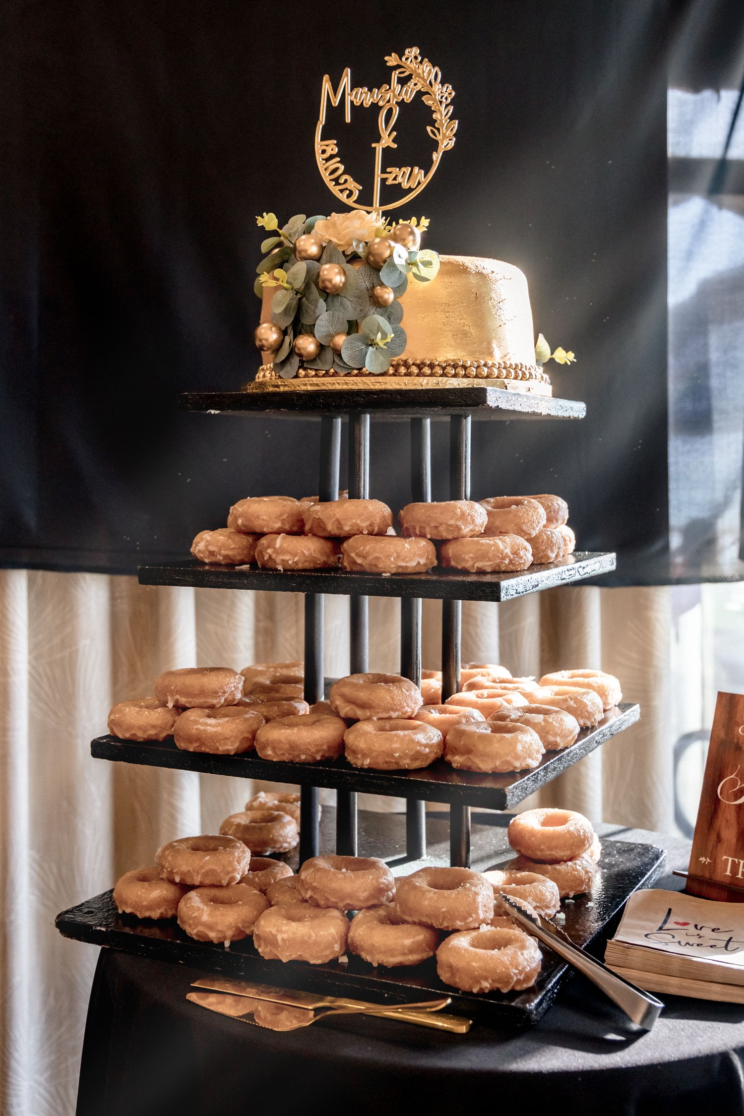 Multi-tiered wedding dessert display featuring glazed donuts and a small cake on top against a dark background.