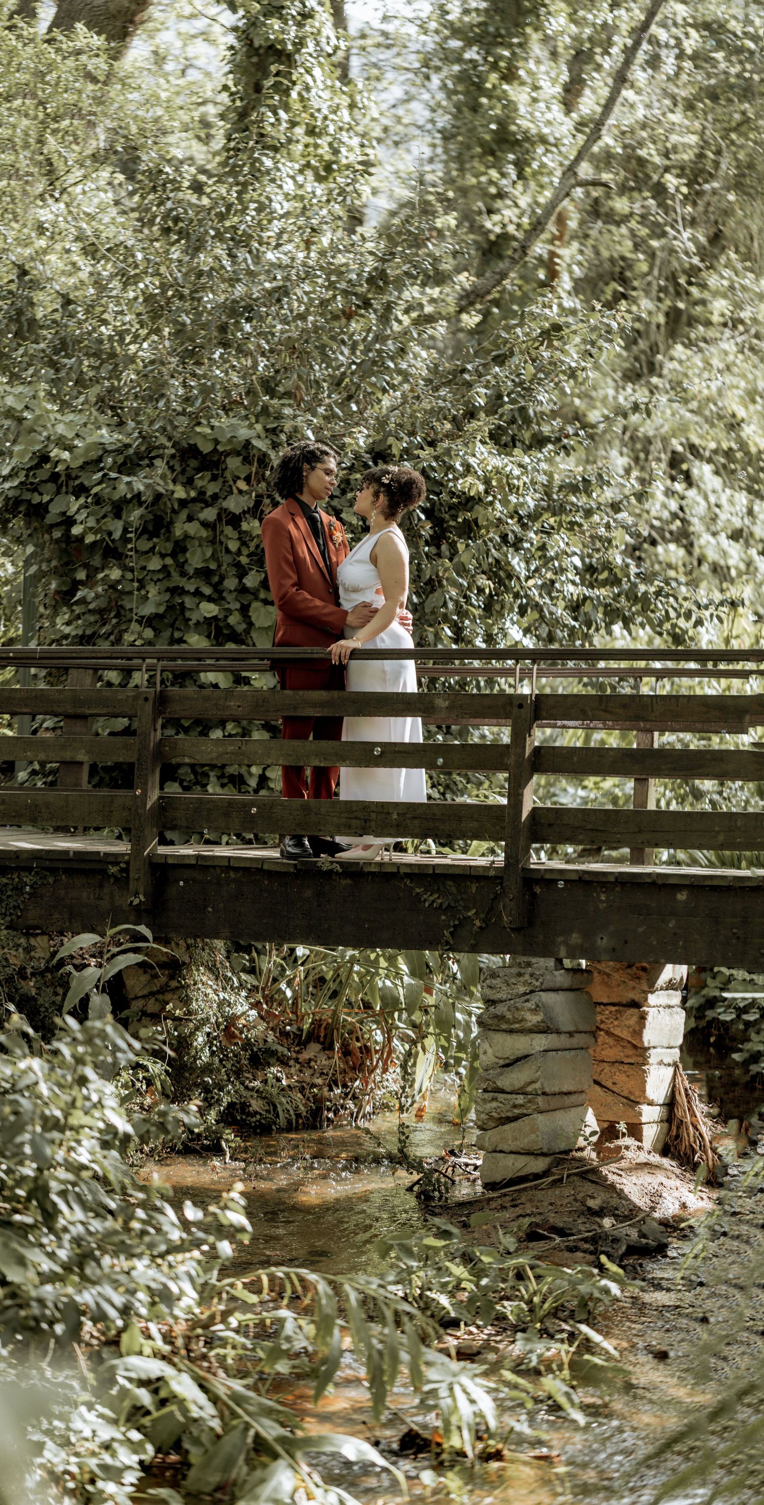 A couple shares an intimate moment together while standing on a wooden footbridge surrounded by lush greenery.