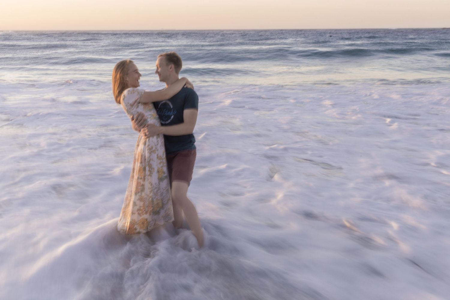 A couple embraces while standing in ocean waves at sunset creating a romantic silhouette against the water.