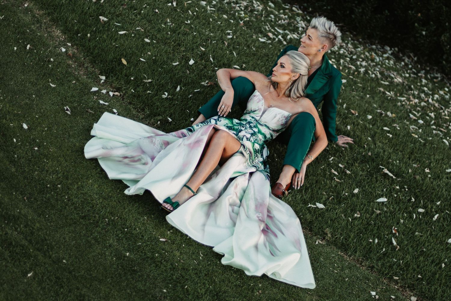 A bride in a white wedding dress sits gracefully on grass covered with scattered white flower petals.