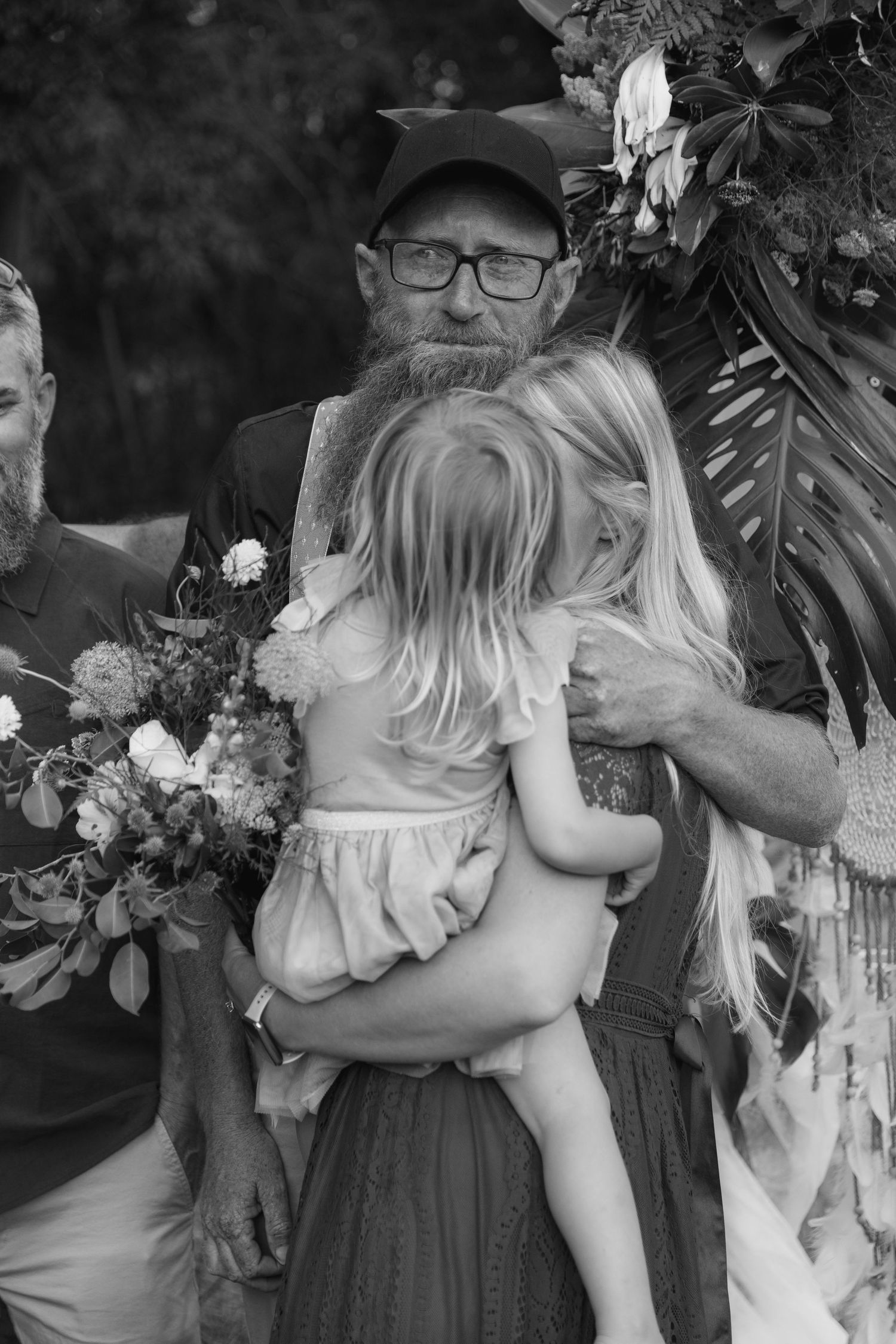 Black and white wedding photo of bearded man in cap hugging young blonde girl, with bouquet and floral arch backdrop.