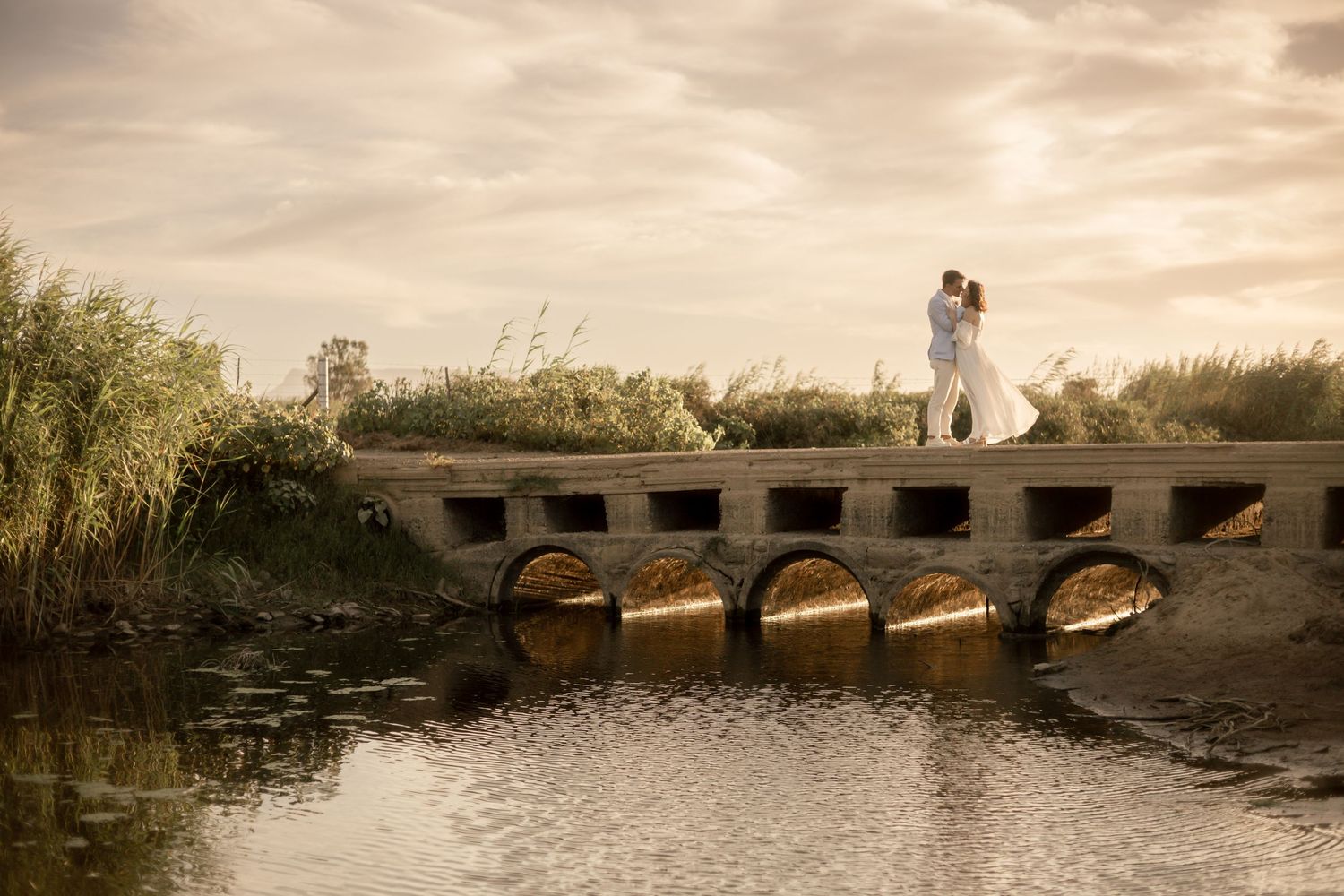 Couple in wedding attire embracing on a stone bridge over calm water at golden hour.