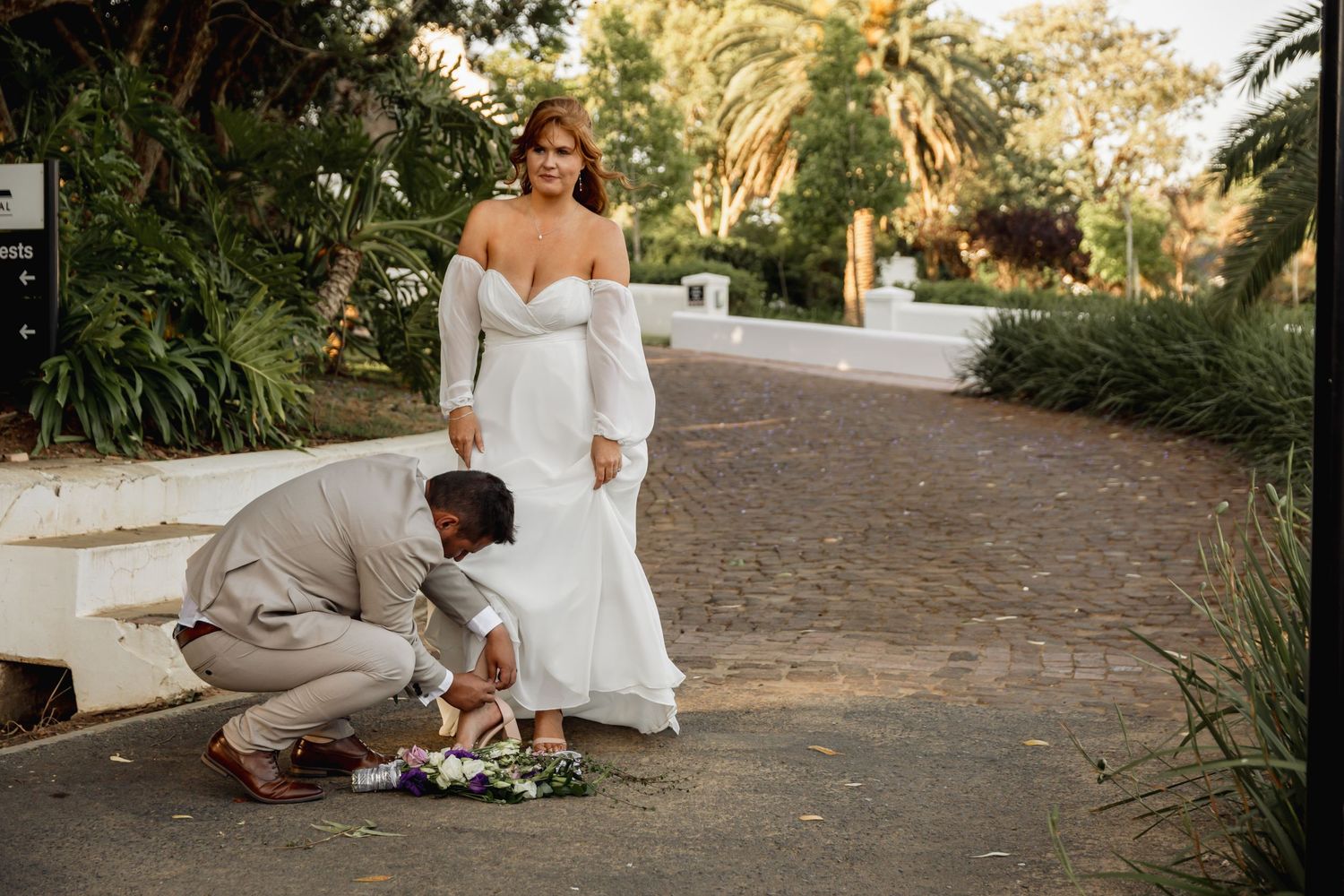 Groom kneeling to arrange bride's wedding bouquet on garden path, bride in elegant white off-shoulder gown.