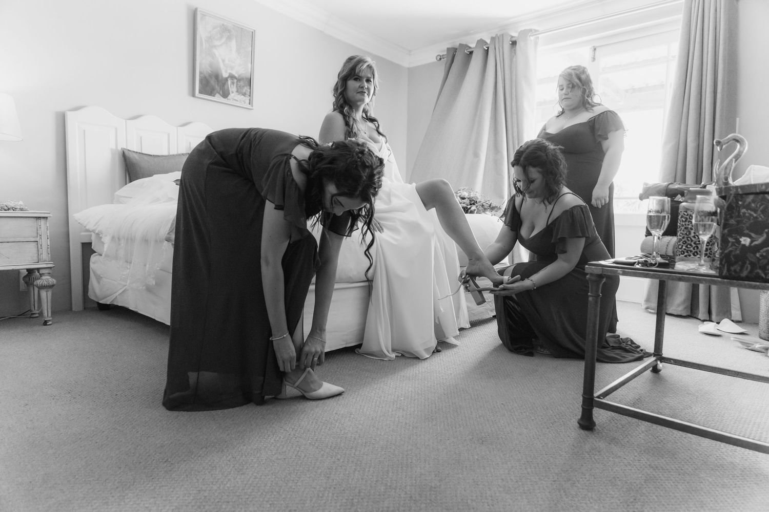 Bridesmaids helping bride with shoes and dress in bedroom during wedding day preparations, black and white photo.