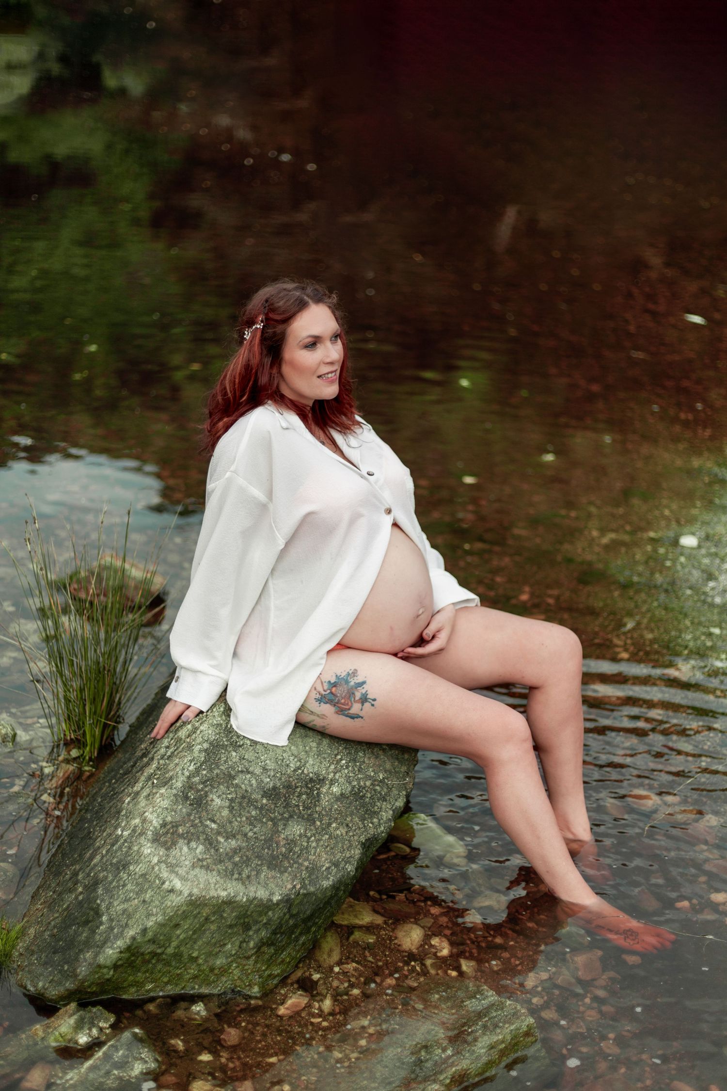 A pregnant woman in a white shirt reclines on a mossy rock by the water's edge, dipping her feet in the stream.
