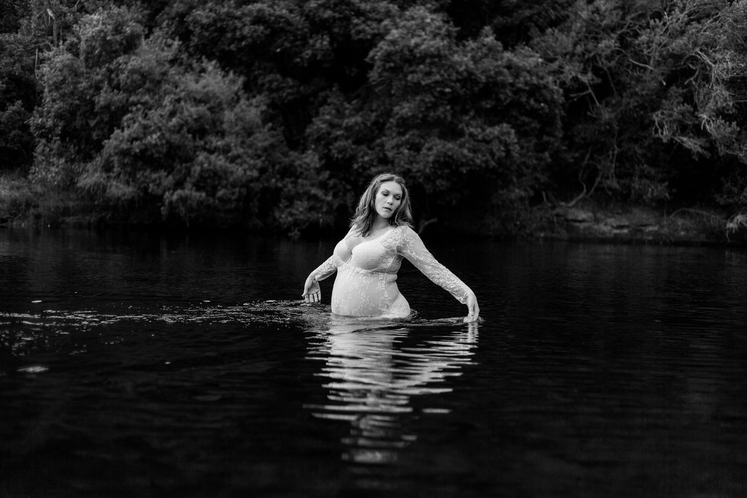 Black and white maternity photo of an expectant mother standing in a serene lake surrounded by trees.