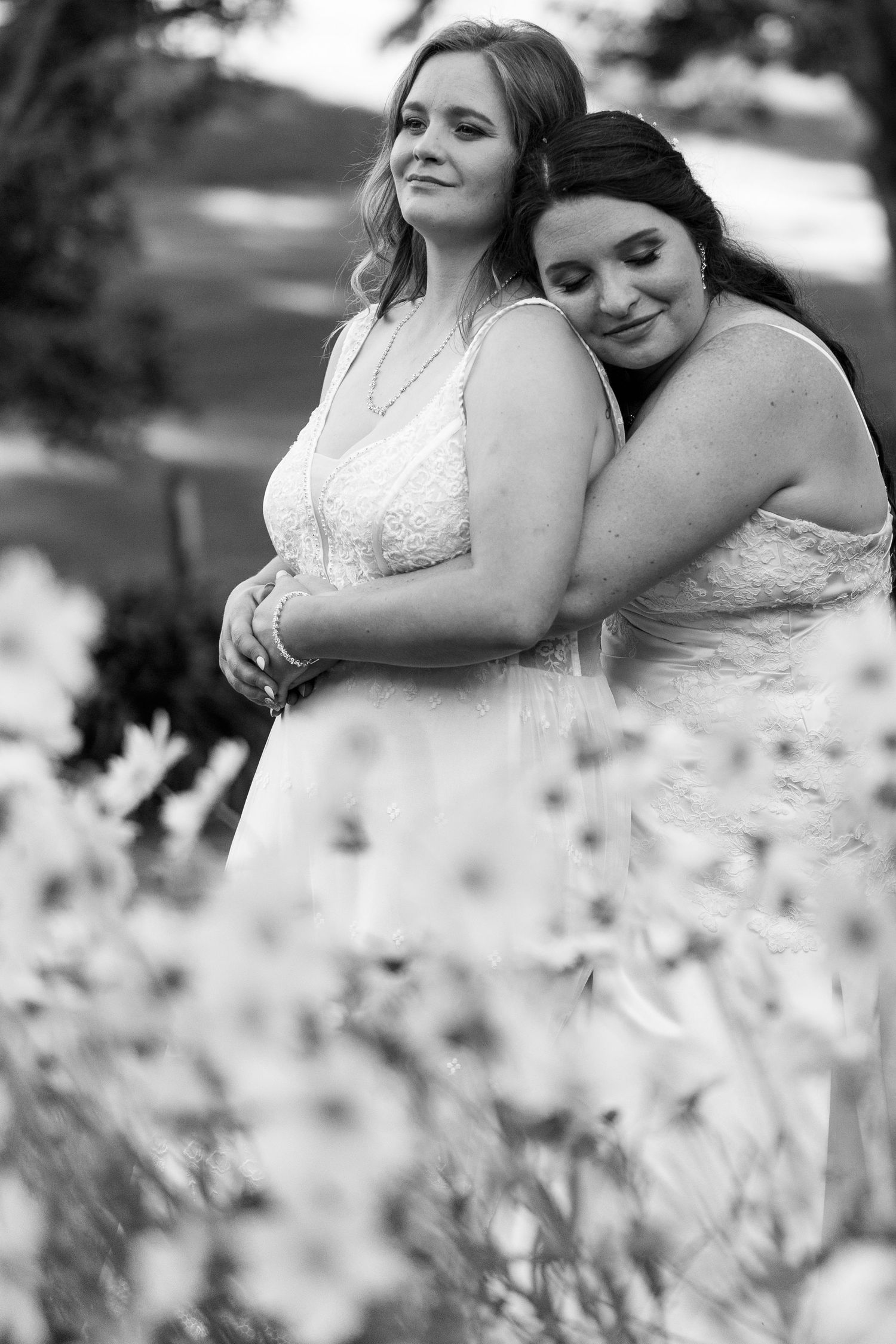 Two people in white dresses embrace in a black and white photo surrounded by wildflowers in a romantic outdoor pose.