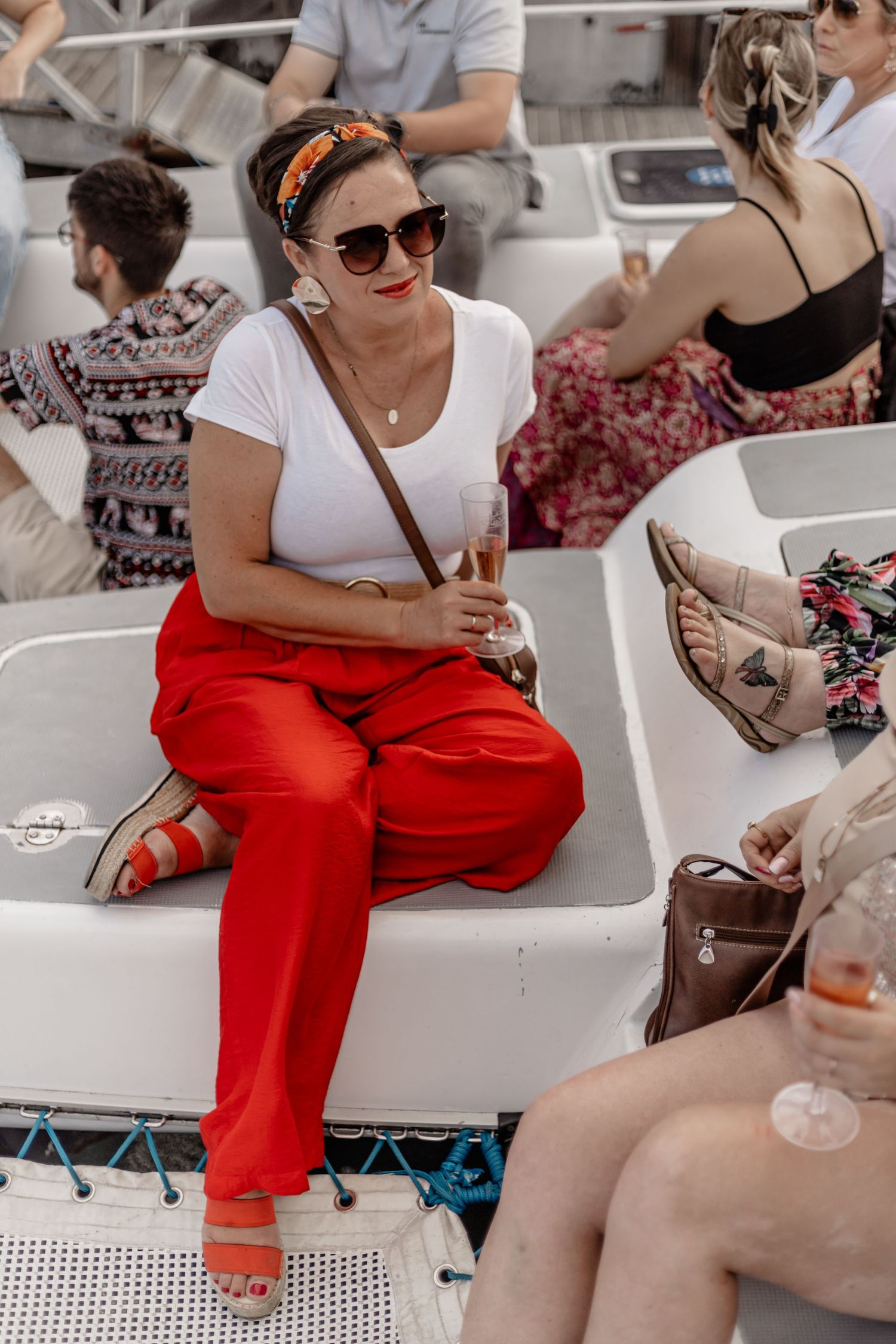 Woman in bright red pants sits casually on white seating while chatting with others on a sunny day.