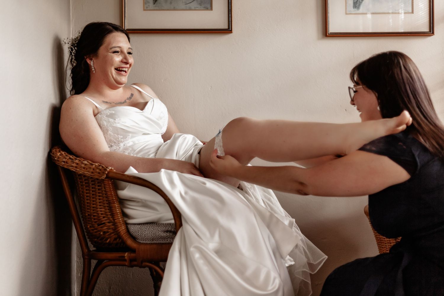 A woman and her bridesmaid get ready for a wedding while wearing white dresses and seated in vintage chairs.