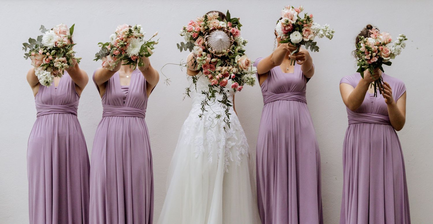 A group of bridesmaids in dusty mauve dresses stand in a line holding floral bouquets at an elegant wedding.
